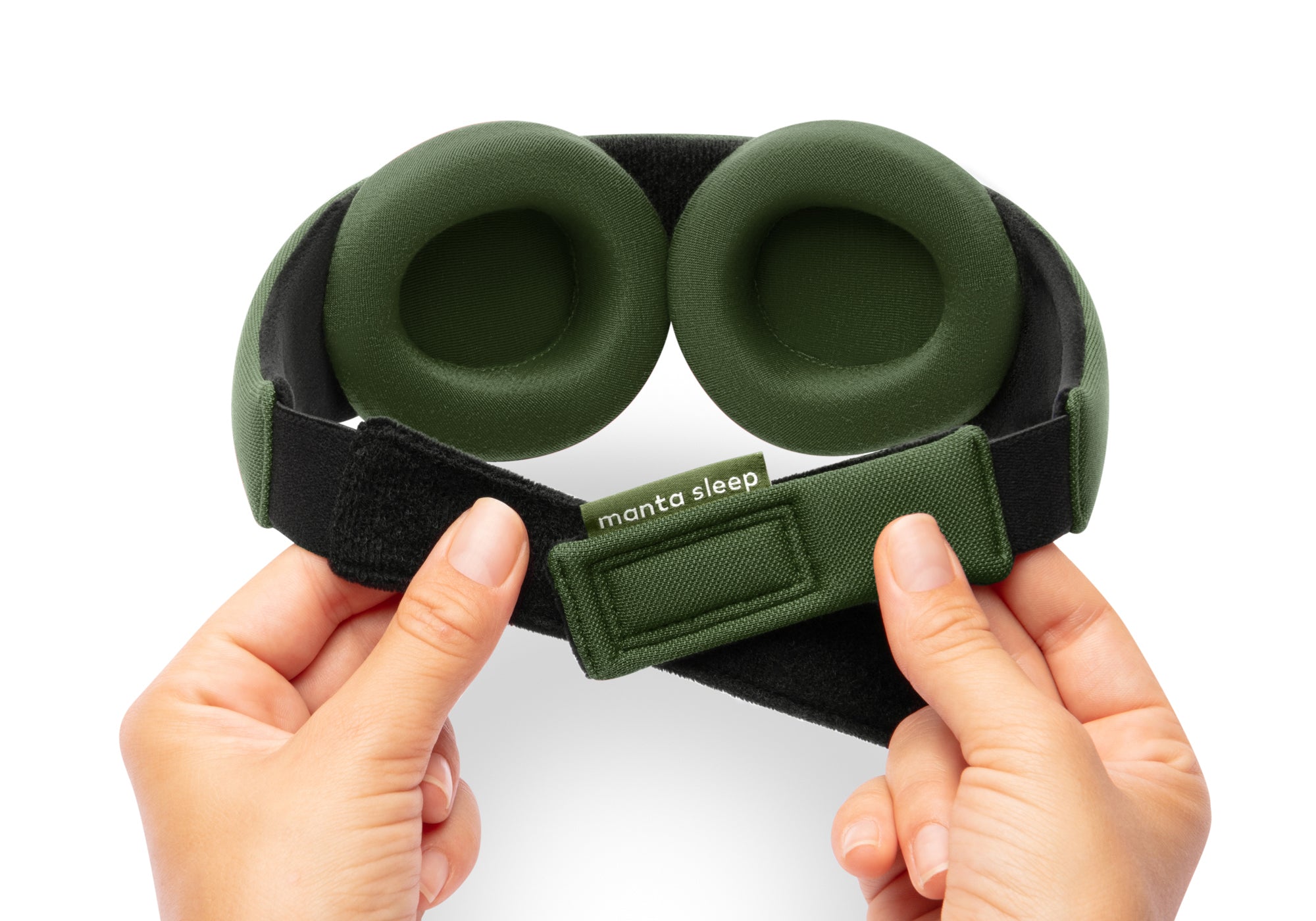 A top-down close-up view of a pair of hands holding the rear hook-and-loop closure of a forest green Manta Sleep Mask against a solid white background, highlighting the adjustable strap alignment and a green fabric tag reading "manta sleep".