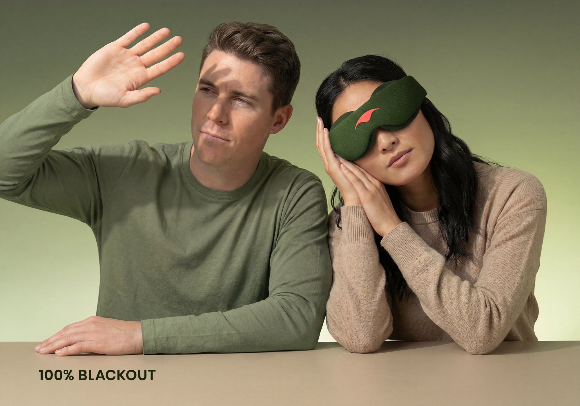 Two people sit side-by-side at a table against a muted green background. The person on the left wears a green long-sleeved shirt and blocks the light with an open hand, while the person on the right wears a tan sweater and rests peacefully while wearing the forest green Manta Sleep Mask. The white text "100% blackout" is in the bottom left corner.