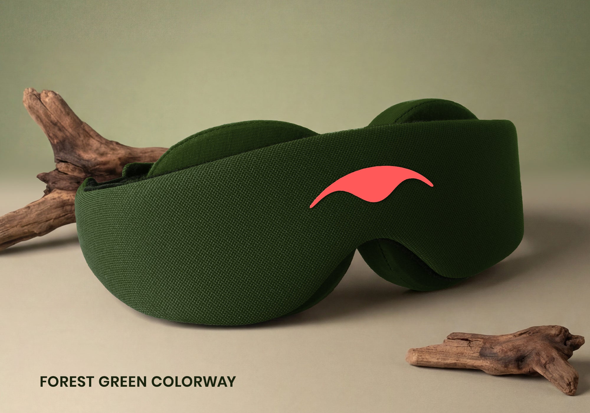 A close-up product shot of the forest green Manta Sleep Mask resting on a light surface alongside a piece of natural driftwood. The smooth coral-red manta ray logo is visible on the front center, and the bold text "Forest green colorway" is displayed in the bottom left corner.