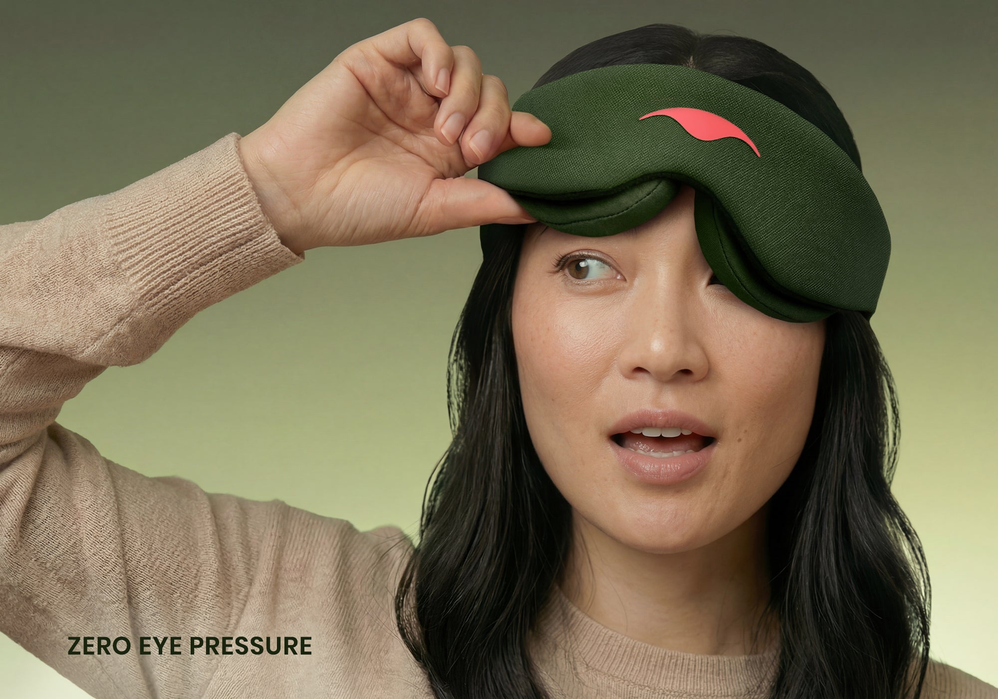 A close-up portrait of a person with dark hair lifting one side of a forest green Manta Sleep Mask away from their eye against a green background. They are wearing a tan sweater, and the white text "Zero eye pressure" is visible in the bottom left corner.