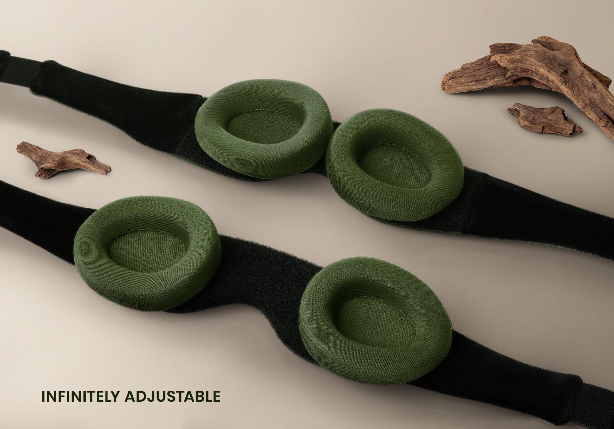 A high-angle studio shot displaying two black sleep mask headbands laid parallel on a light beige surface, highlighting the adjustable, circular forest green eyecups attached to them. Decorative pieces of natural driftwood are arranged around the masks, with the text "Infinitely adjustable" at the bottom left.