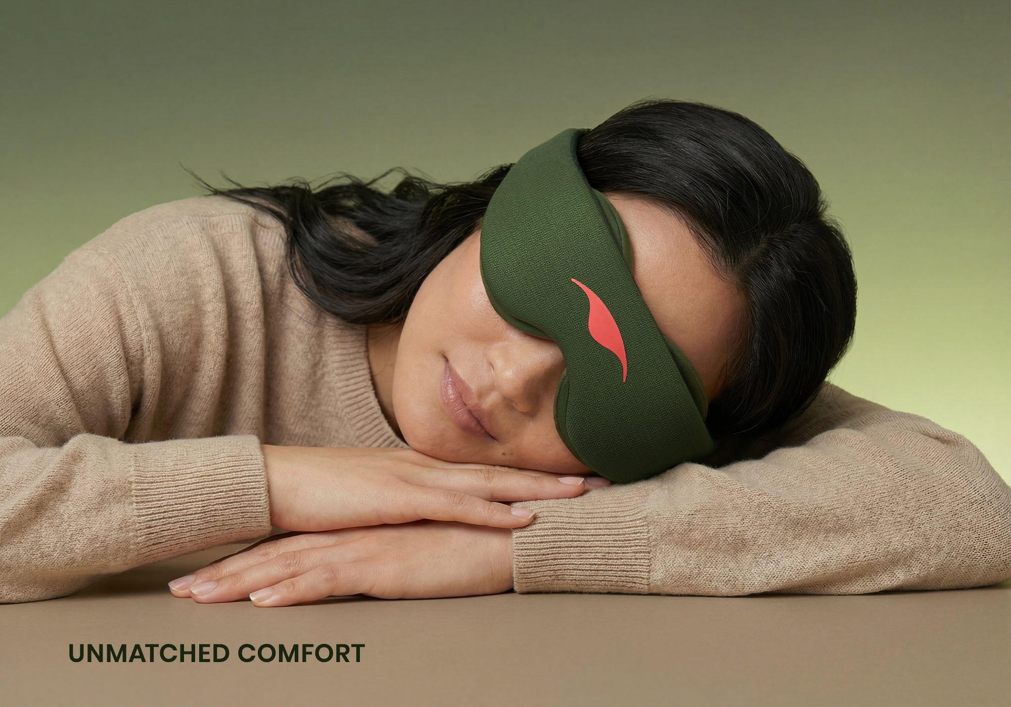 A person with dark hair rests their head on their crossed arms on a flat surface, appearing to sleep comfortably while wearing a forest green Manta Sleep Mask. They are wearing a tan knit sweater against a muted green gradient background, with the white text "Unmatched comfort" displayed in the bottom left corner.