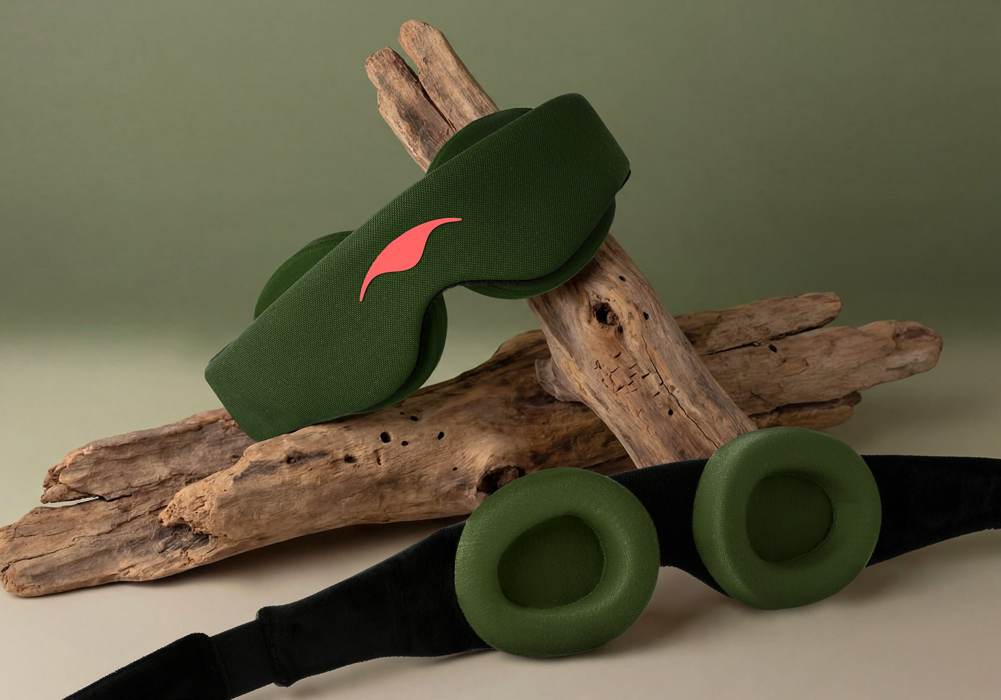 A studio product display showing two Forest Green Manta Sleep Masks arranged with pieces of natural driftwood against a muted green background. One mask rests angled on top of the wood to show the front coral-red logo, while the other mask lies flat in the foreground, showcasing its two thick, circular, and contoured green interior eyecups.