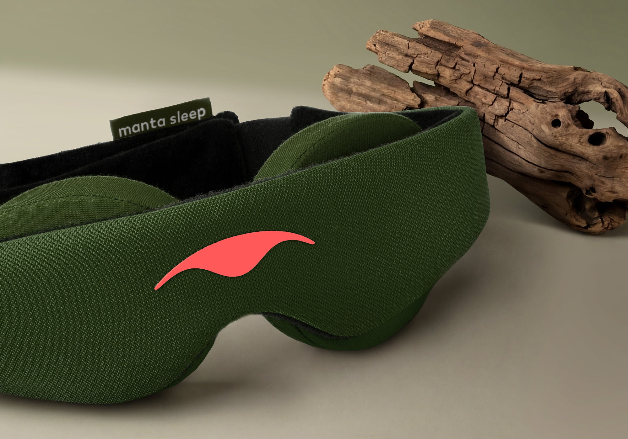 A close-up studio shot of the Forest Green Manta Sleep Mask resting on a light beige surface next to a piece of textured, natural driftwood. The mask highlights its fine mesh texture, a vibrant coral-red printed logo on the front, and a dark green brand label reading "manta sleep" along the top edge.