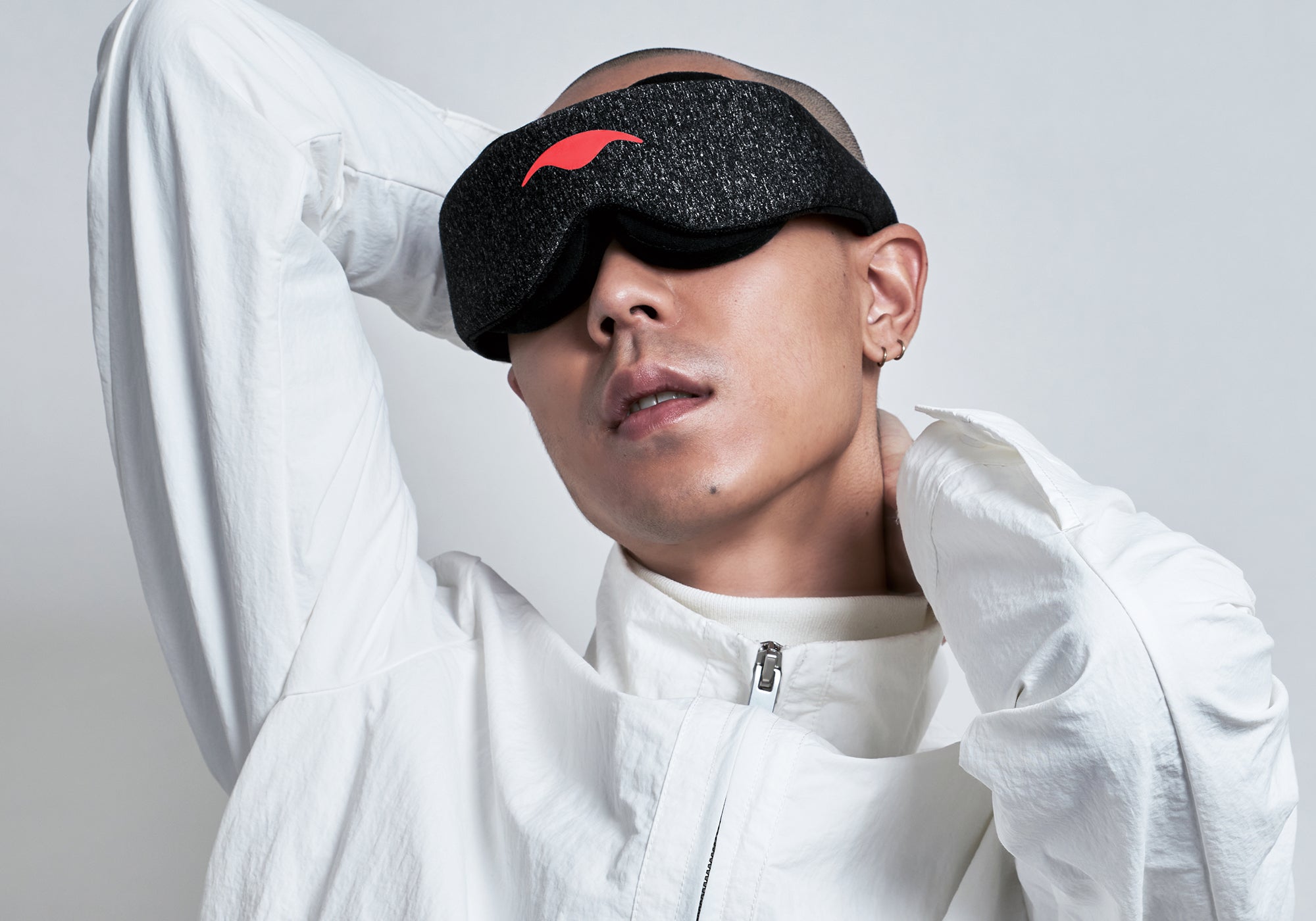 A studio portrait of a person with a shaved head wearing a dark gray heathered Carbon Black Manta Sleep Mask over their eyes. They are wearing a zippered white jacket and have their hands positioned behind their head and neck against a light gray backdrop.