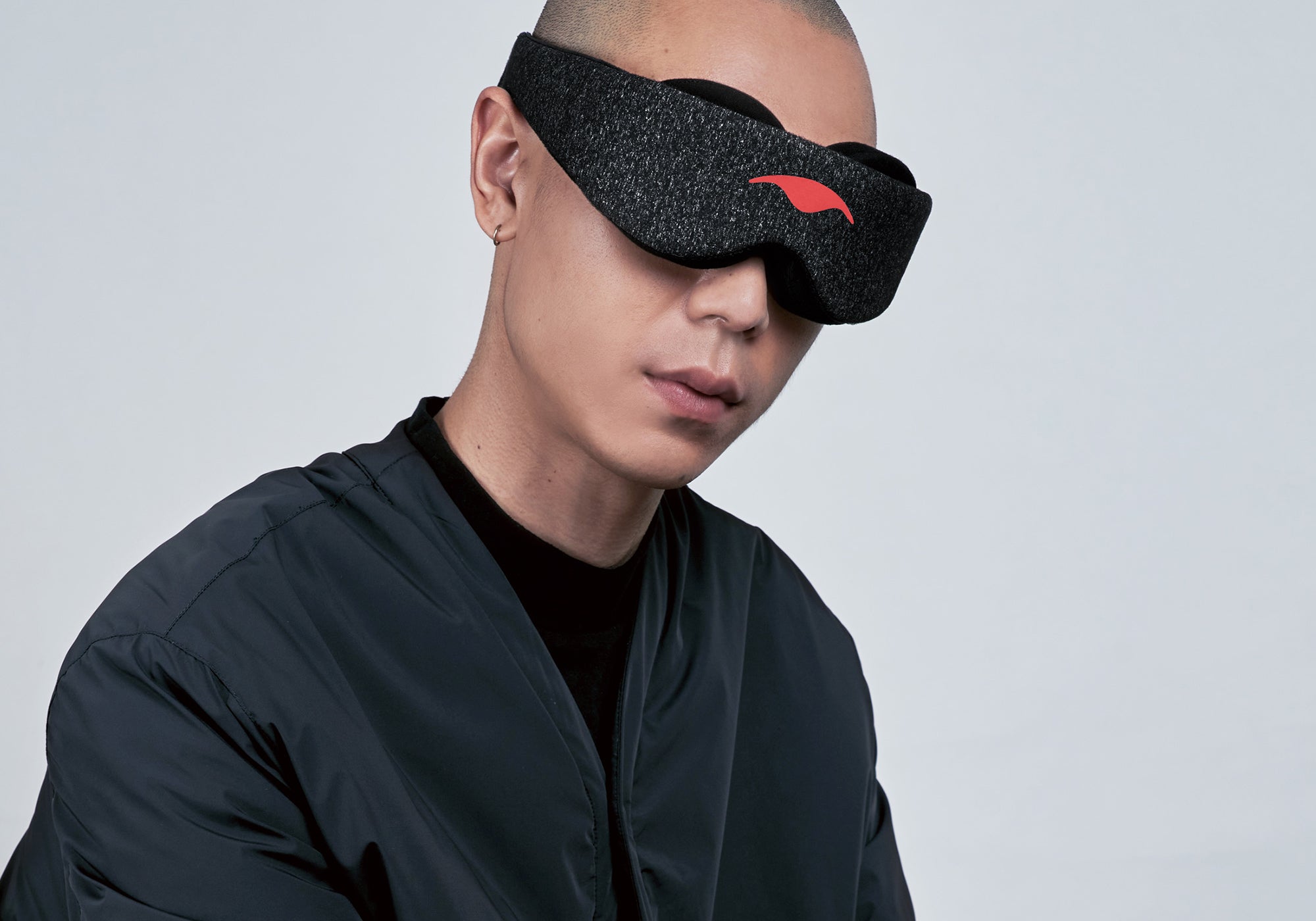 A studio portrait showing a person with a shaved head from the chest up, angled slightly to the side against a neutral light gray background. They are wearing a dark gray speckled Carbon Black Manta Sleep Mask over their eyes and a dark charcoal zippered jacket over a black shirt.