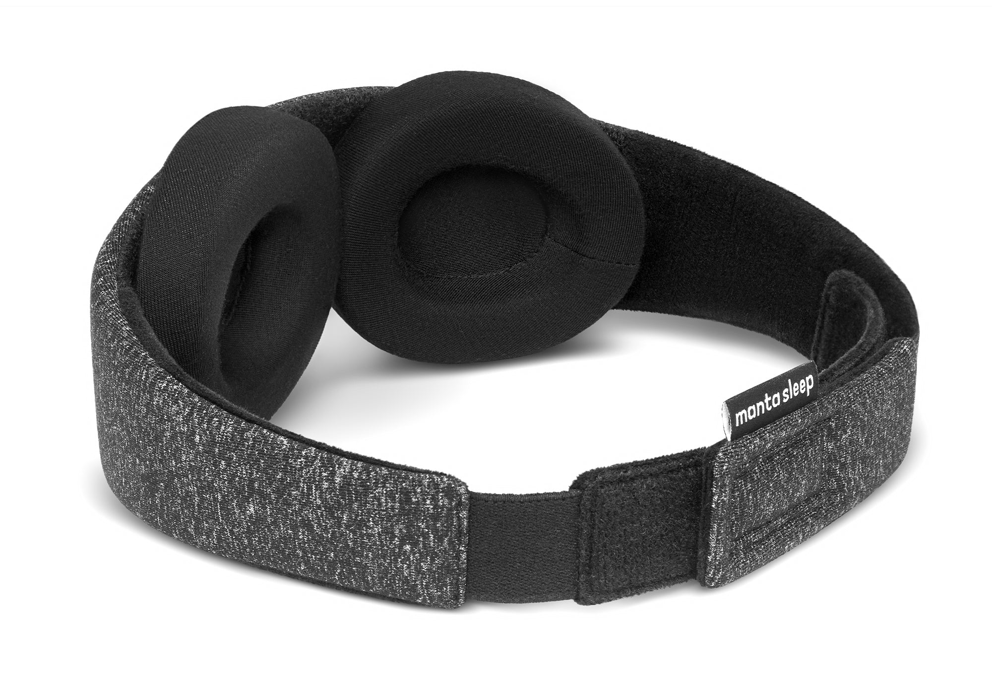 A three-quarter rear perspective of the Carbon Black Manta Sleep Mask isolated on a white background. This view showcases the dark gray heathered exterior strap, the black plush inner lining, the adjustable hook-and-loop closure, and a small black brand label reading "manta sleep".