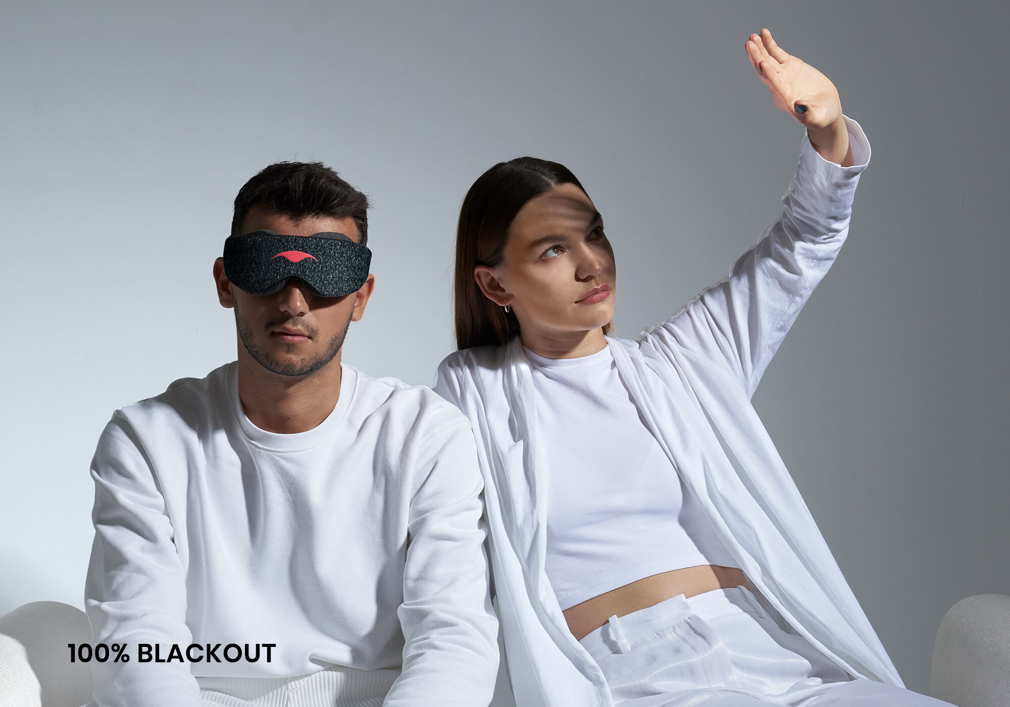 Two people sit side-by-side against a light gray studio background. The person on the left is wearing a white sweatshirt and the Carbon Black Manta Sleep Mask, while the person on the right wears a white top and holds a hand up to shield their eyes from a shadow cast across their face. The white text "100% blackout" is in the bottom left corner.