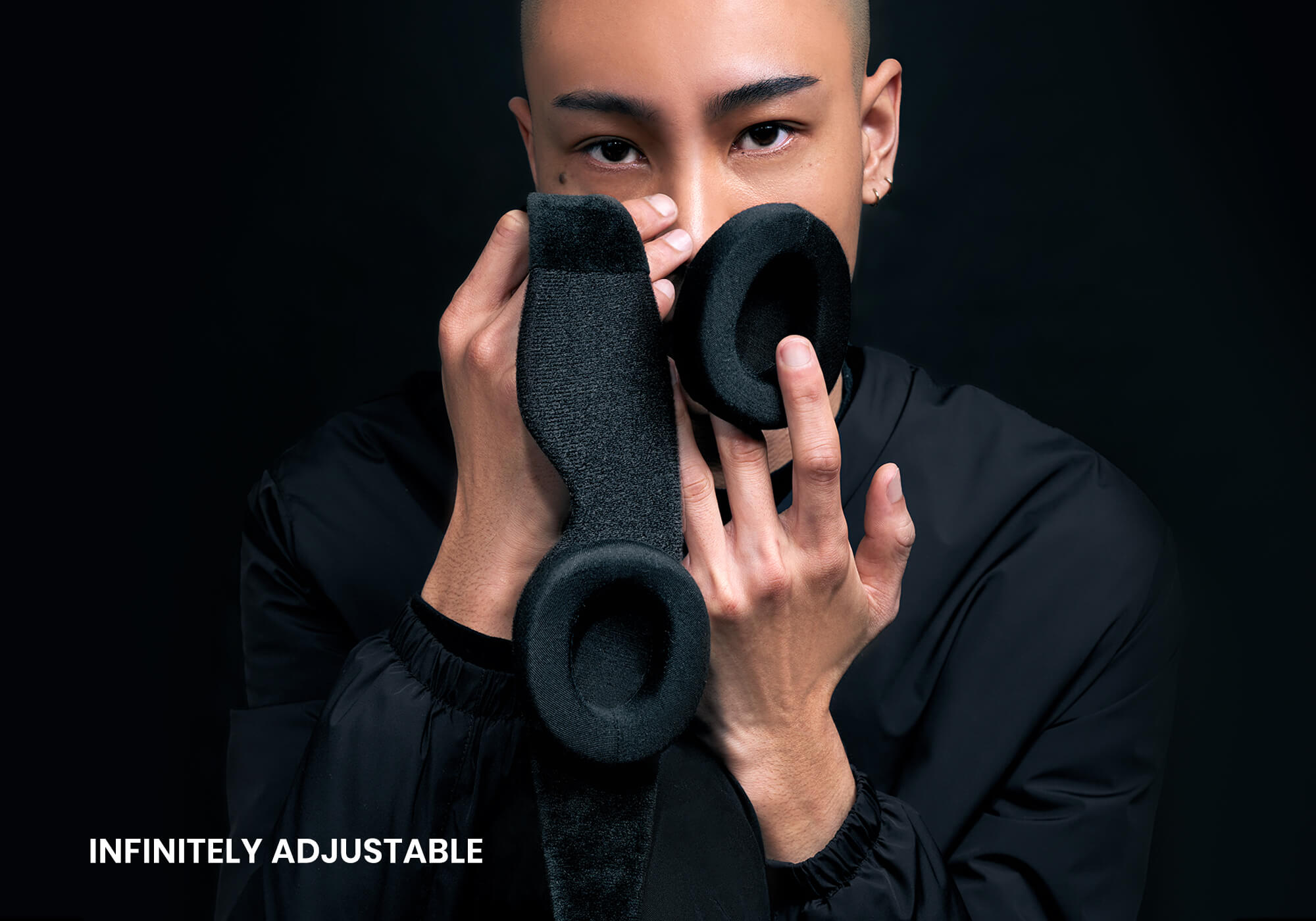 A close-up portrait of a person holding the detached black eyecup components of the sleep mask up to their face against a solid black background. The white text "Infinitely adjustable" is displayed in the lower left corner.