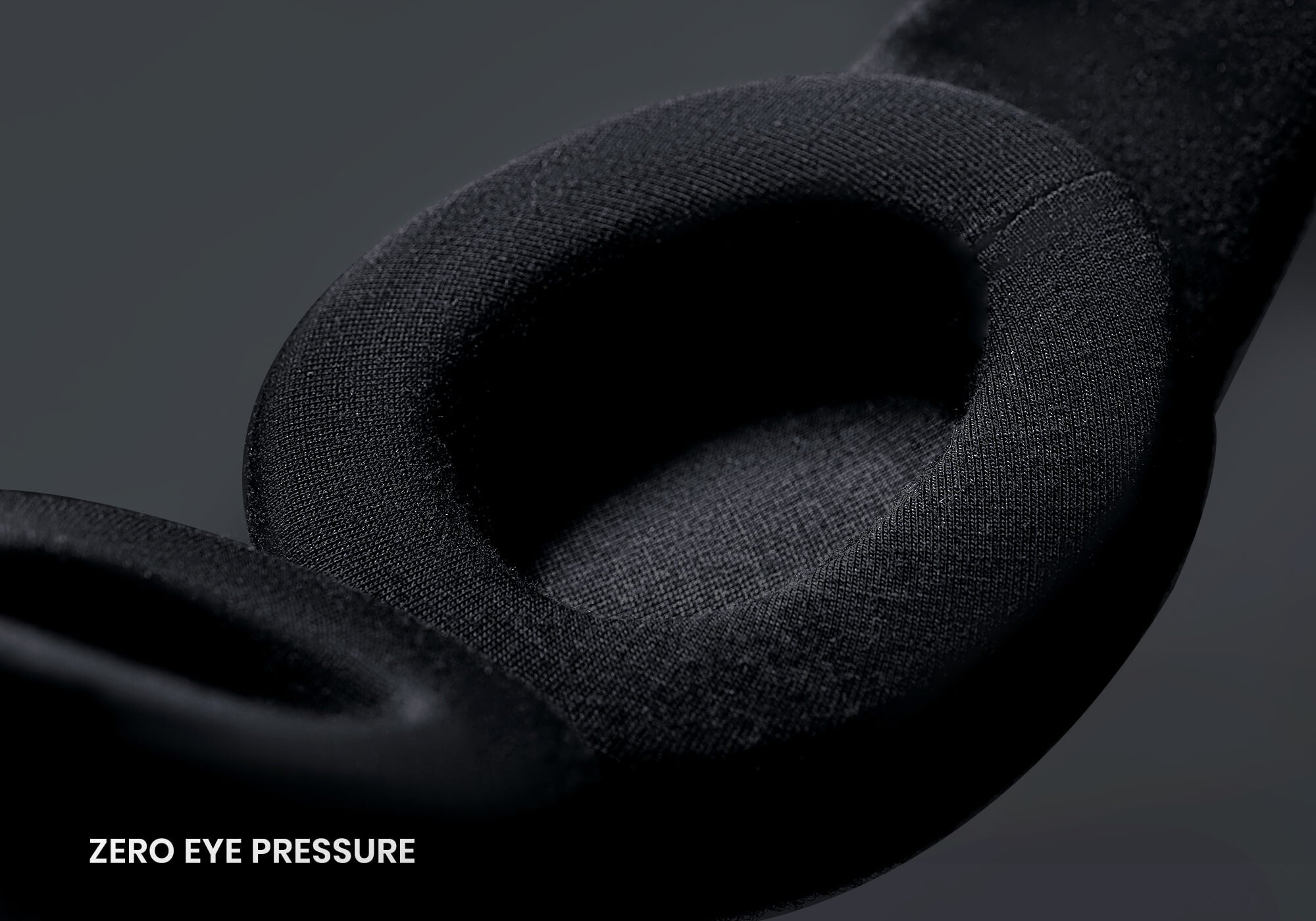 A macro, high-angle view focusing on the interior of a black sleep mask. It features a thick, contoured, oval-shaped eyecup covered in a smooth black knit material, with the white text "Zero eye pressure" located in the bottom left corner.