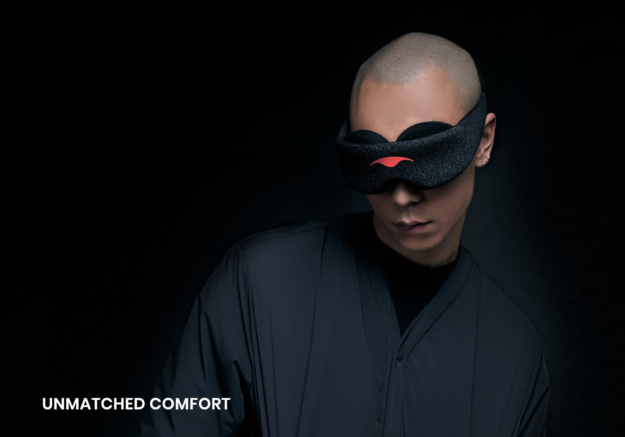 A dramatic studio portrait of a person wearing the Carbon Black Manta Sleep Mask, looking down slightly against a pitch-black background. They are wearing a dark charcoal-colored shirt, and the white text "Unmatched comfort" is visible in the bottom left corner.