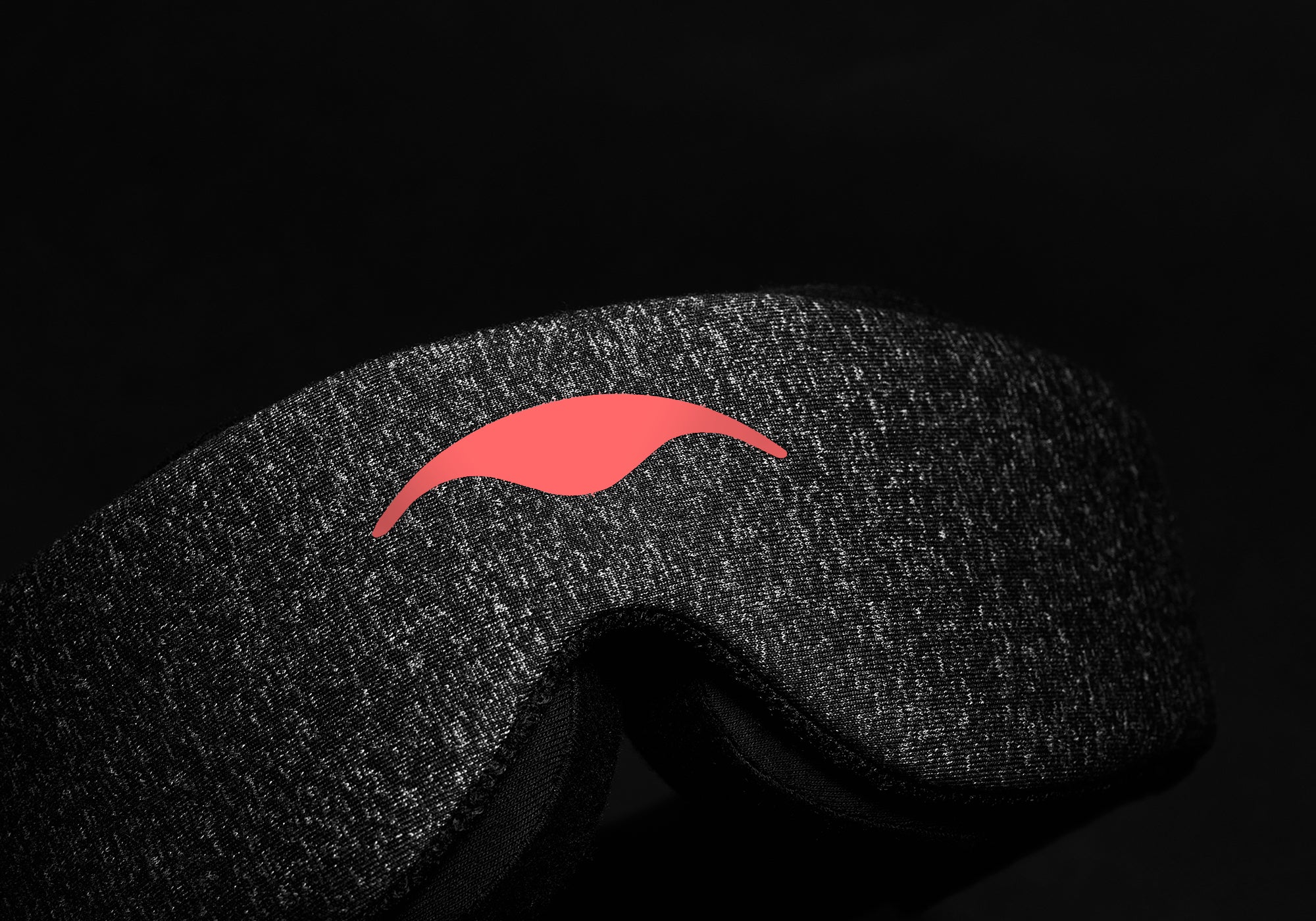 A close-up shot focusing on the textured dark gray heather fabric of the Carbon Black Manta Sleep Mask against a solid black background. The smooth coral-red Manta Sleep logo is prominently displayed in the center.