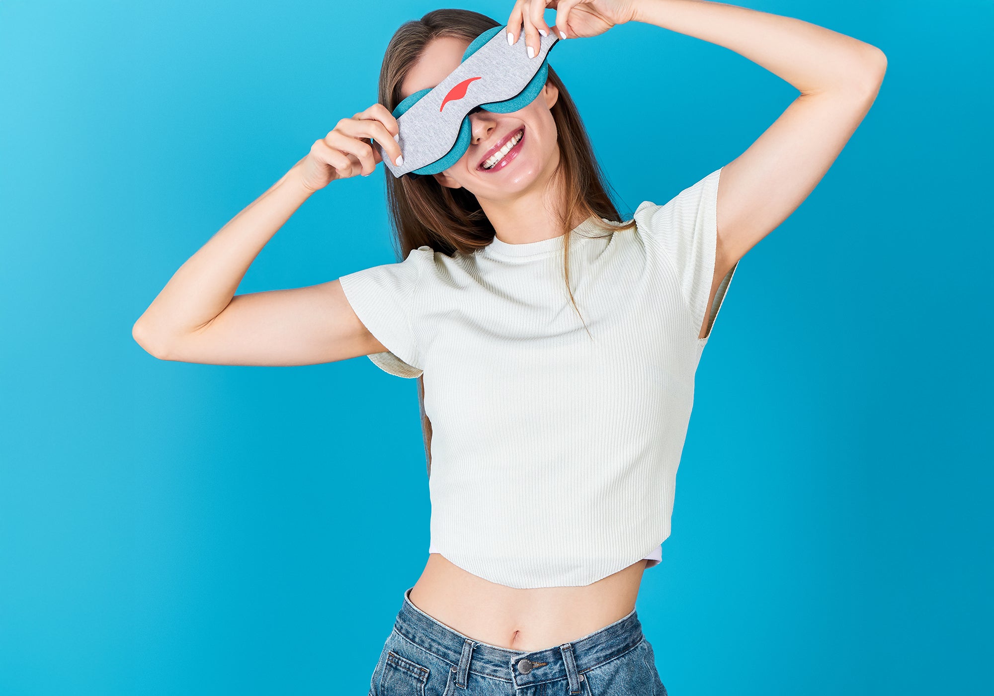 A smiling woman holding a grey Manta COOL Sleep Mask with a coral logo over her eyes, showing the blue-teal eye cups underneath, on a light blue background.