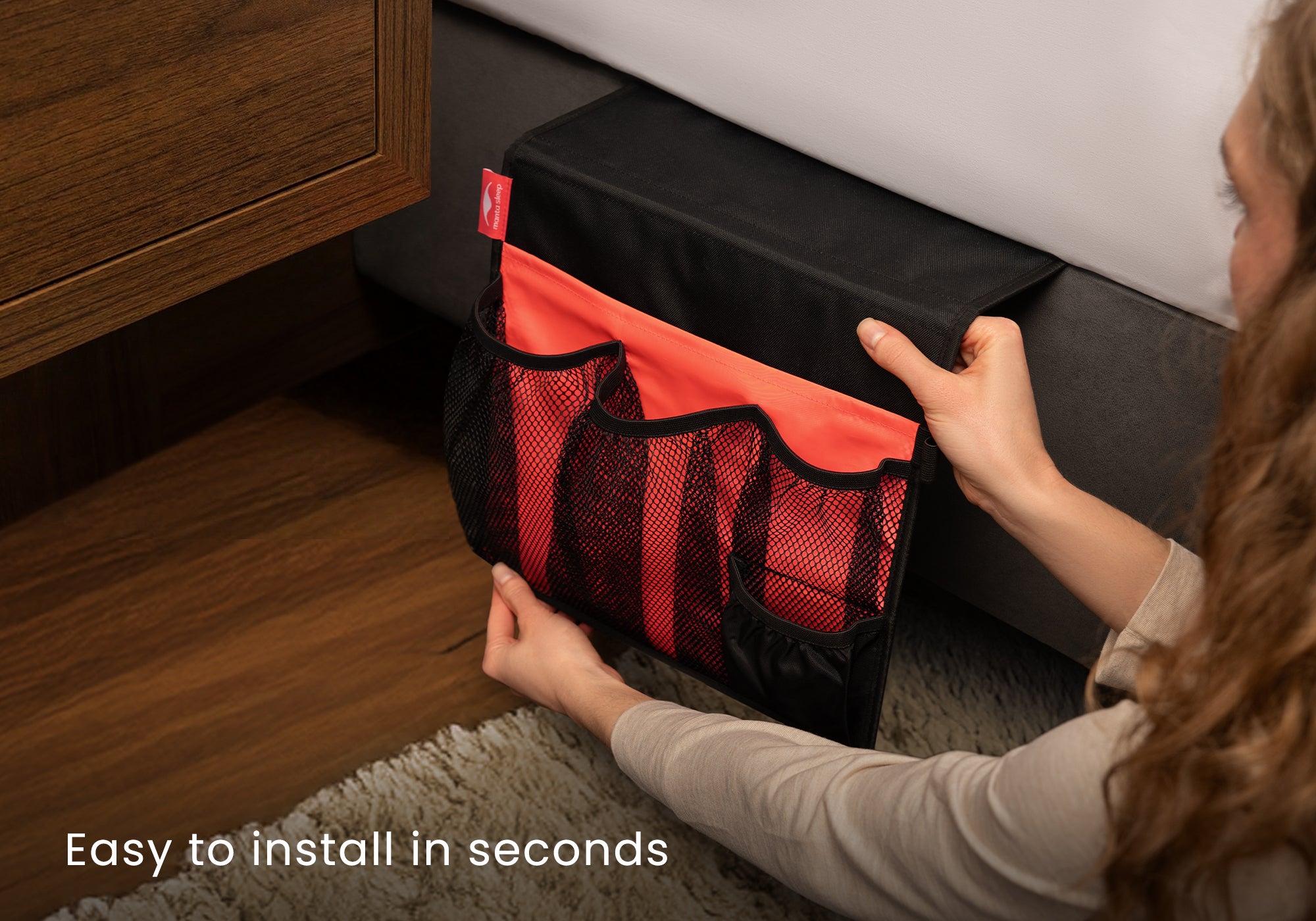 Hands inserting a black and coral Manta Bedside Caddy between a mattress and bed frame. Text overlay reads "Easy to install in seconds".