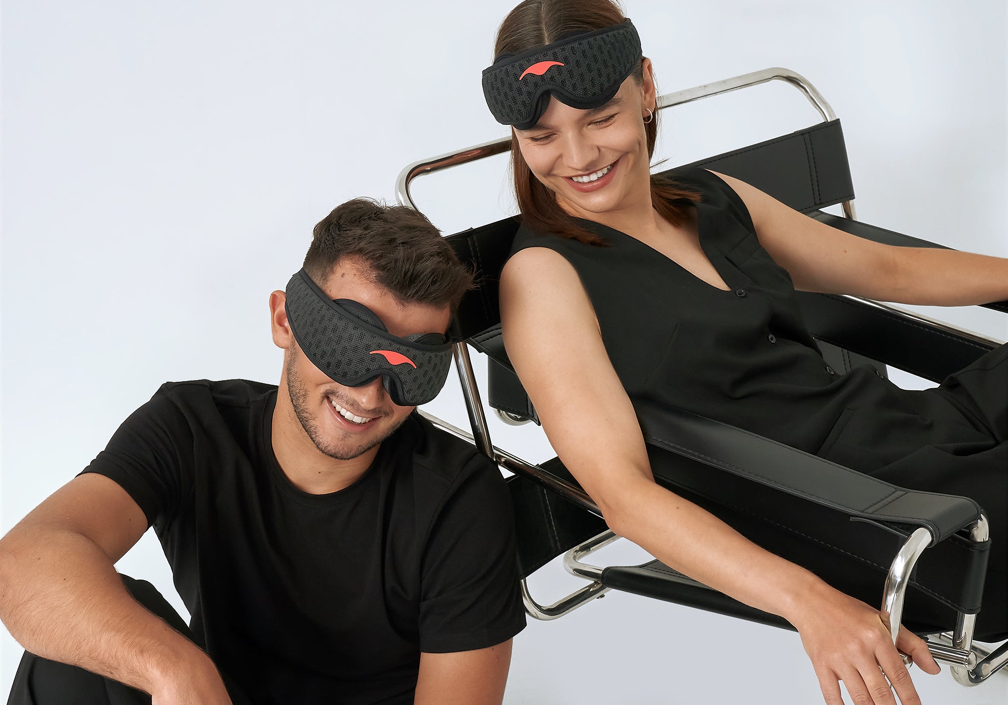 A smiling man and woman pose in black attire with Carbon Black Manta Sleep Mask PROs on their eyes and forehead, sitting next to a modern chair.