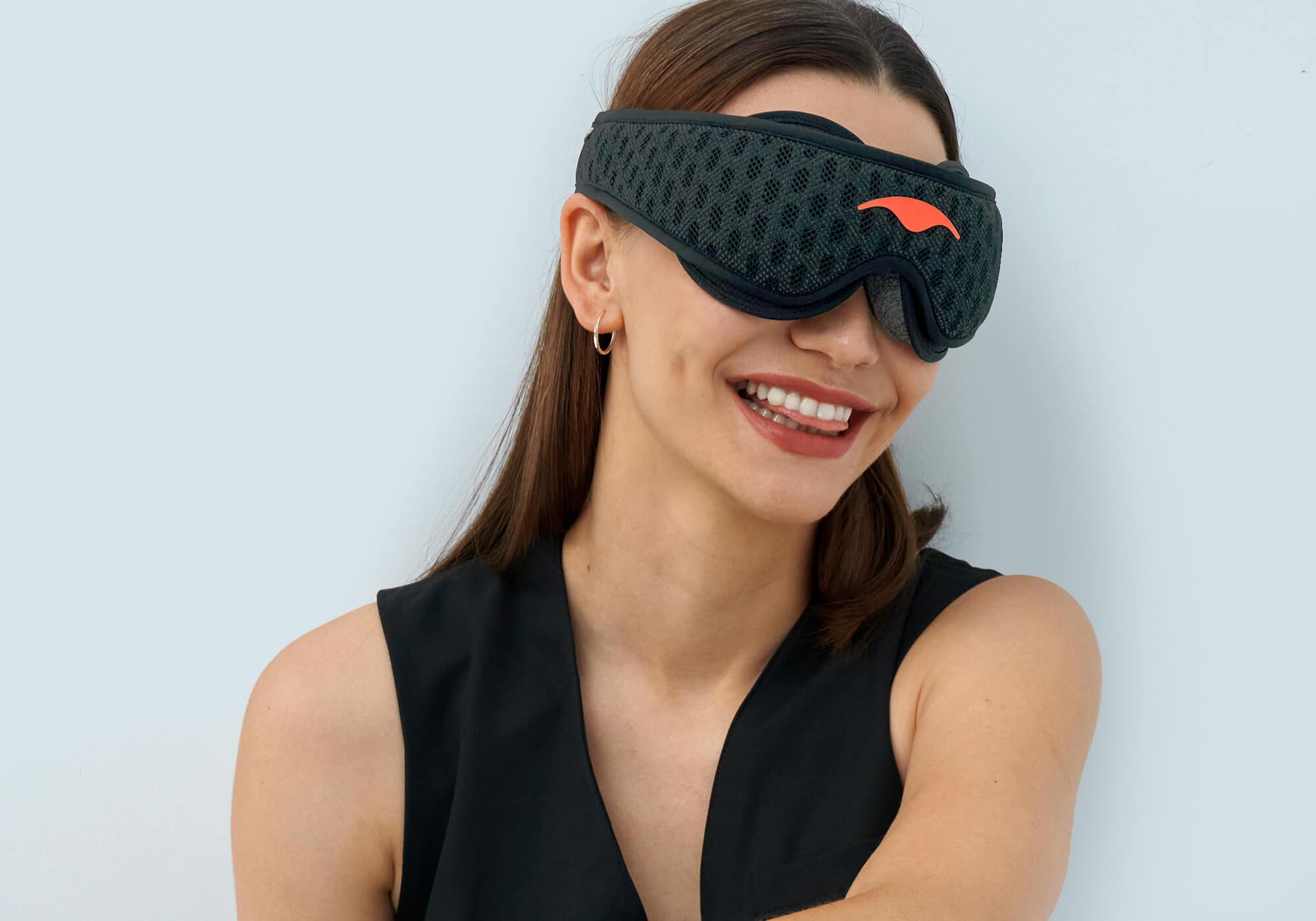 Close-up of a smiling woman wearing a Carbon Black Manta Sleep Mask PRO with a vibrant coral Manta Sleep logo against a light background.