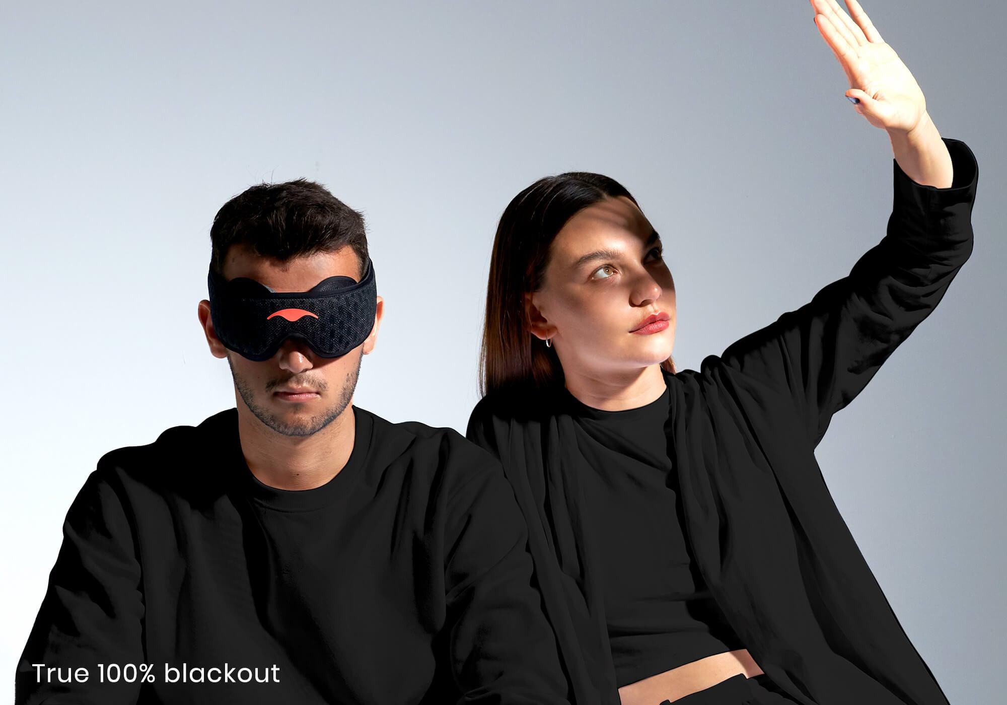 Two people under bright light; one wears a Carbon Black Manta Sleep Mask PRO, while the other shields their eyes, with caption "True 100% blackout".