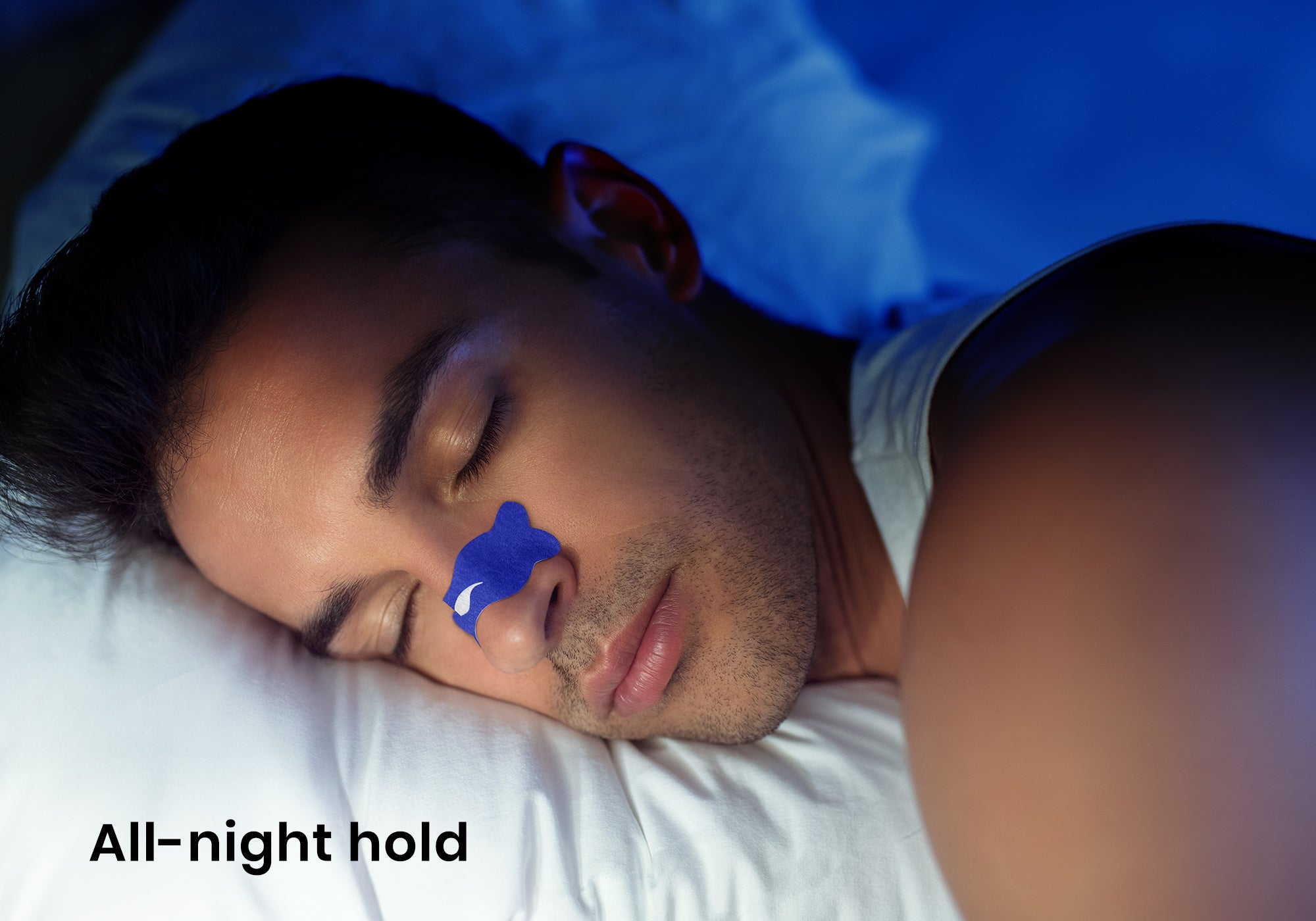 Close-up of a man sleeping with a blue Manta nasal strip on his nose under blue lighting. Text overlay: "All-night hold".