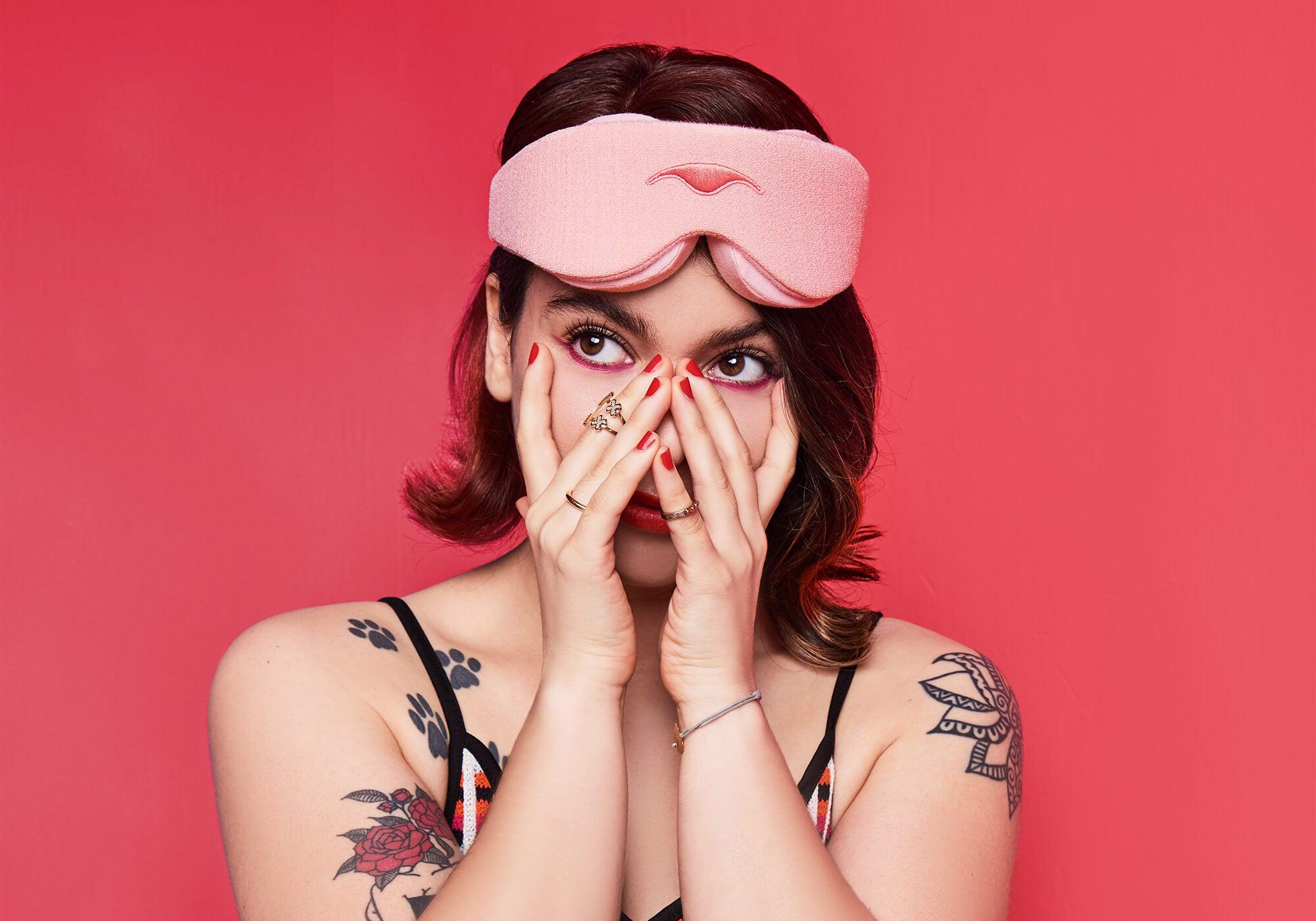 A studio portrait of a person with short brown hair looking upward, framed against a solid dark pink background. They are wearing a plush, light pink Manta Sleep Mask pushed up on their forehead and covering their nose with their hands, showing off red nail polish, rings, and arm tattoos.