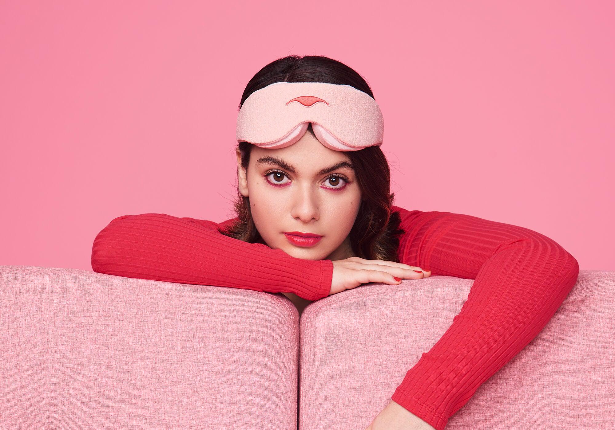 A studio portrait of a person with dark hair wearing a light pink Manta Sleep Mask on their forehead, leaning their arms over the back of a pink sofa against a solid pink background. They are wearing a long-sleeved red ribbed top, pink eyeshadow, and red lipstick while looking directly forward.