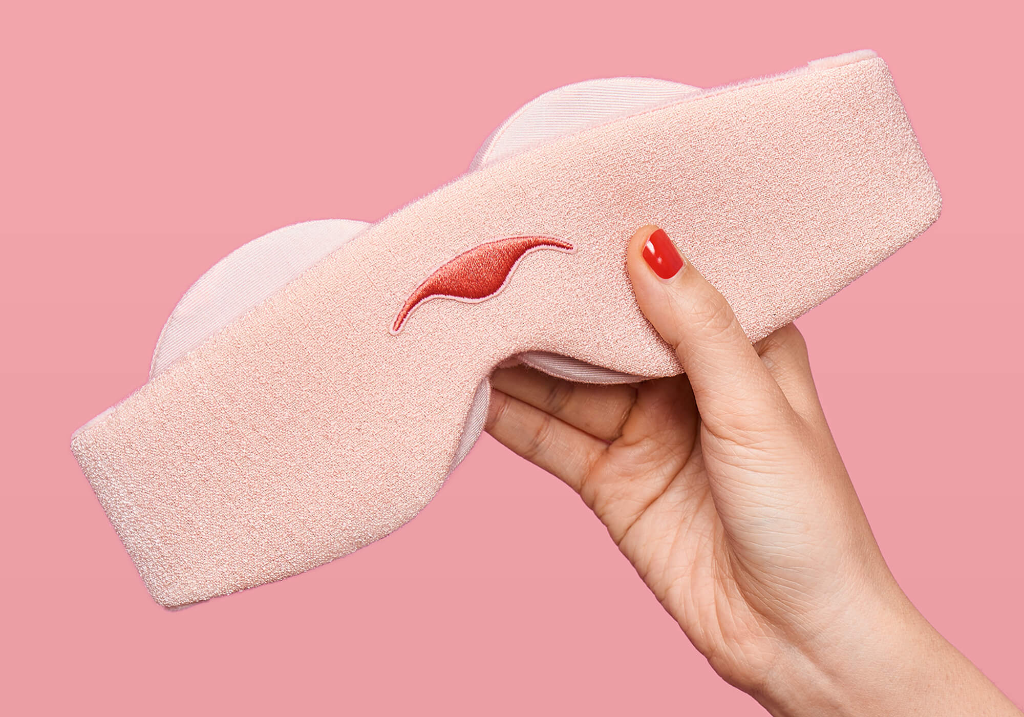 A close-up view of a hand with bright red nail polish holding the Sunrise Pink Manta Sleep Mask diagonally against a solid pink background, showing the embroidered darker pink Manta Sleep logo on the front and the edges of the light pink padded eyecups underneath.