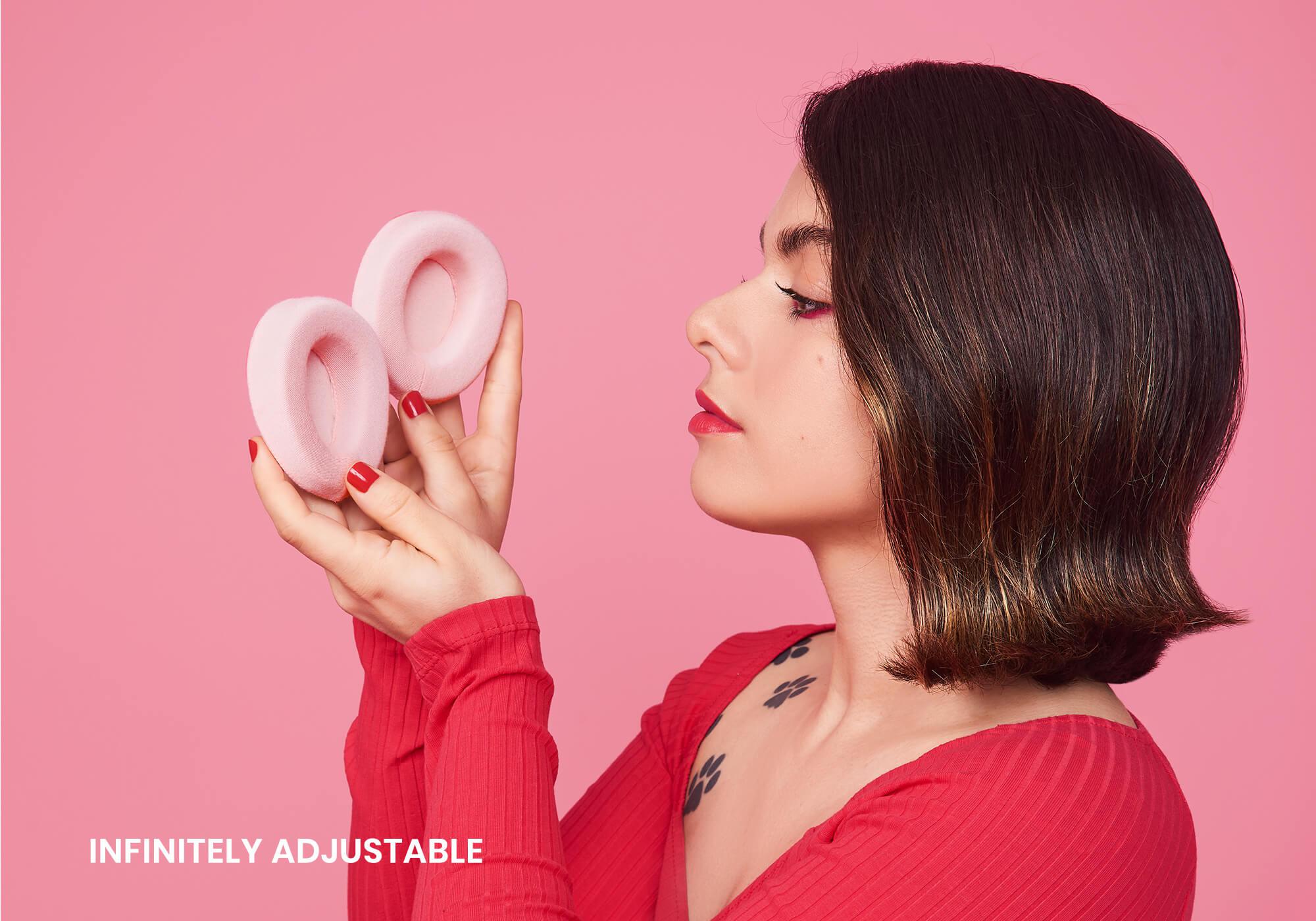 A side profile portrait of a person with dark hair and red lipstick looking at two detachable, plush pink eyecups held in their hands. The background is a solid pink color, and the white text "Infinitely adjustable" is positioned in the bottom left corner.