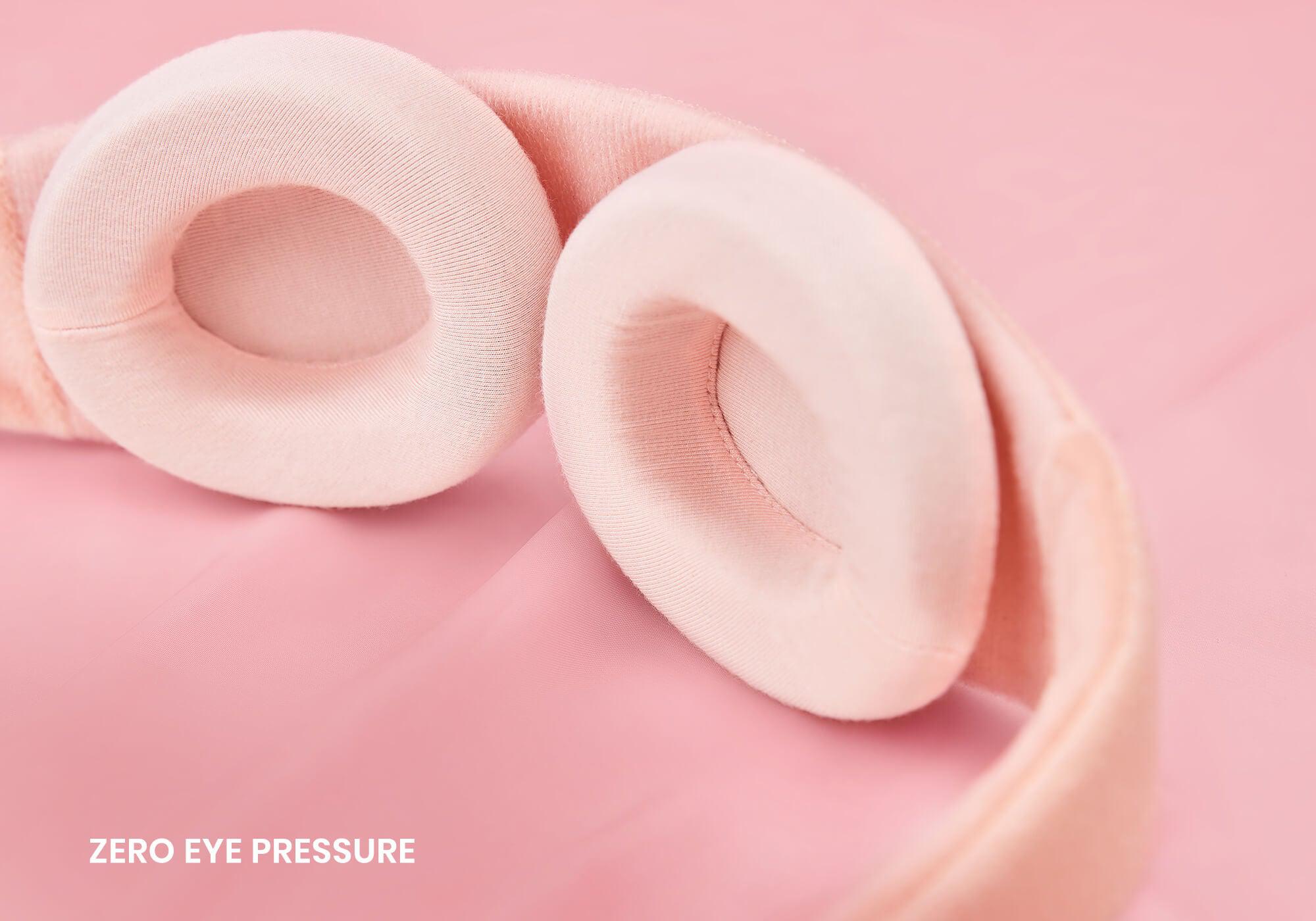A close-up, high-angle view focusing on the interior of a light pink sleep mask resting on a soft pink fabric surface. Two thick, oval-shaped cushioned eyecups in a smooth pink knit material are prominently featured, with the white text "Zero eye pressure" visible in the bottom left corner.
