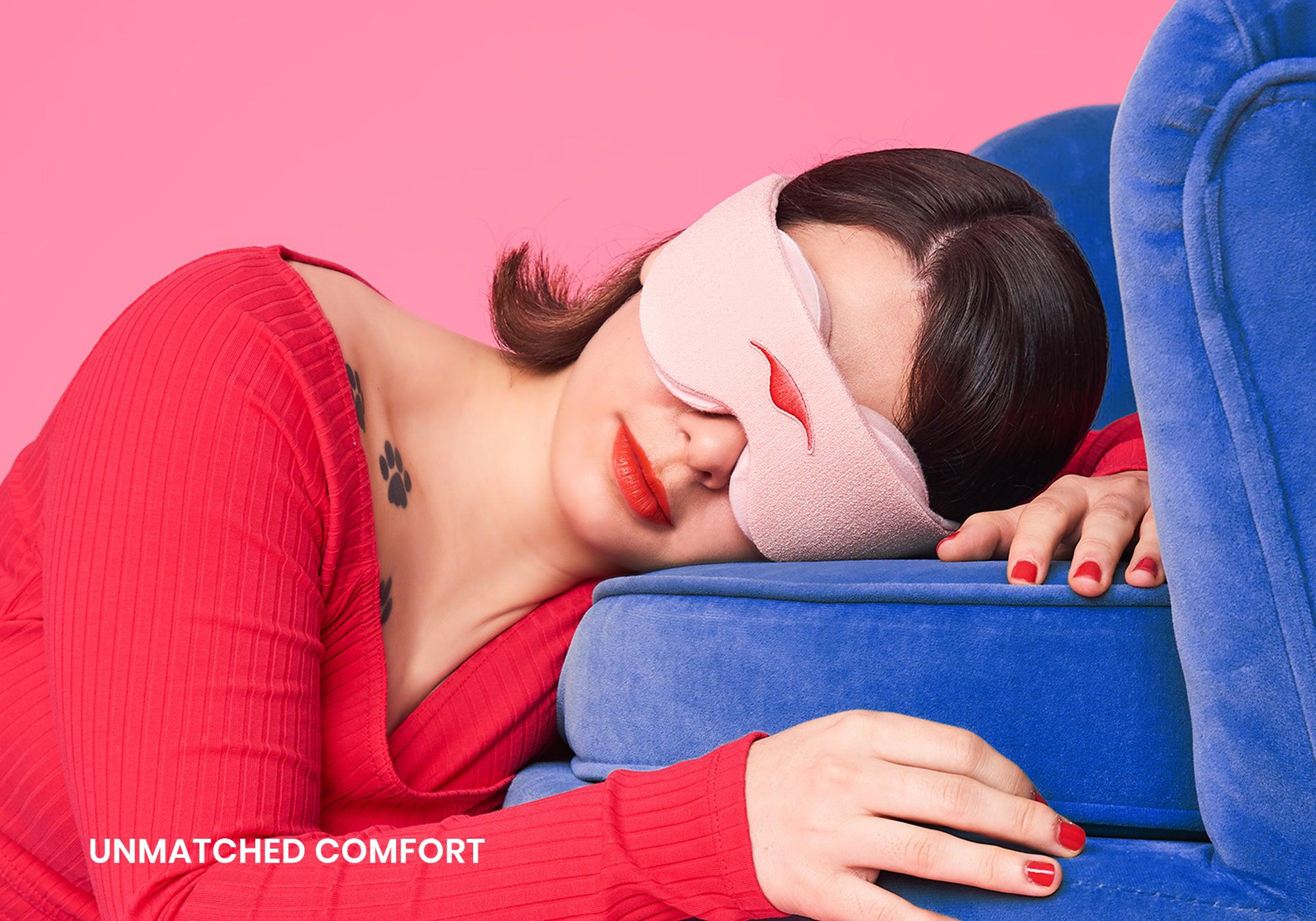 A person in a red ribbed long-sleeve top rests their head comfortably against a plush, bright blue velvet armchair. They are wearing a light pink Manta Sleep Mask that fully covers their eyes, set against a pink background, with the white text "Unmatched comfort" in the bottom left corner.