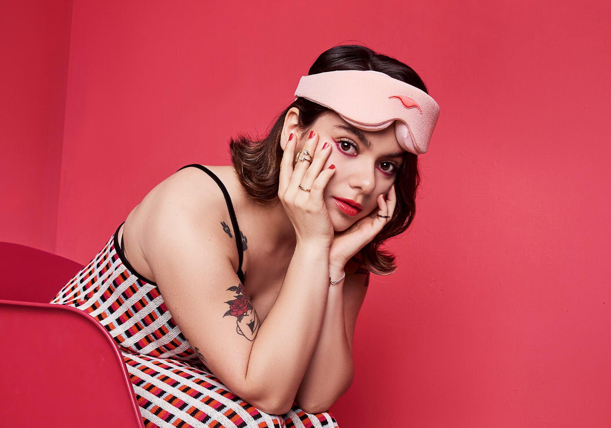 A person with short dark hair rests their chin on their hands while leaning forward in a bright pink chair against a matching pink background. They are wearing a light pink Manta Sleep Mask pushed onto their forehead, a patterned sleeveless top, and bright red lipstick.