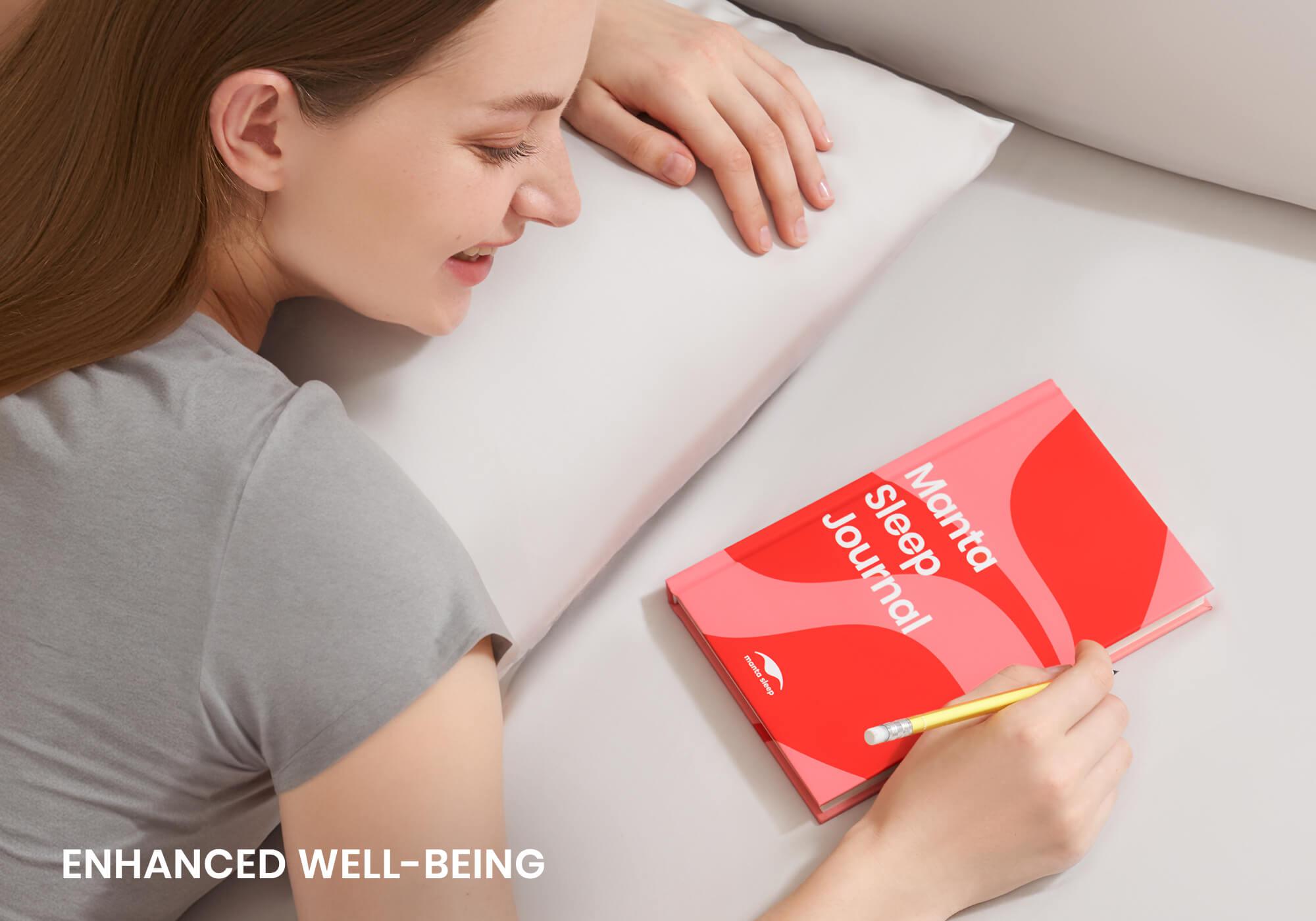 A smiling woman resting her head on a white pillow next to a red Manta Sleep Journal on the bed. Overlay text reads "Enhanced well-being".