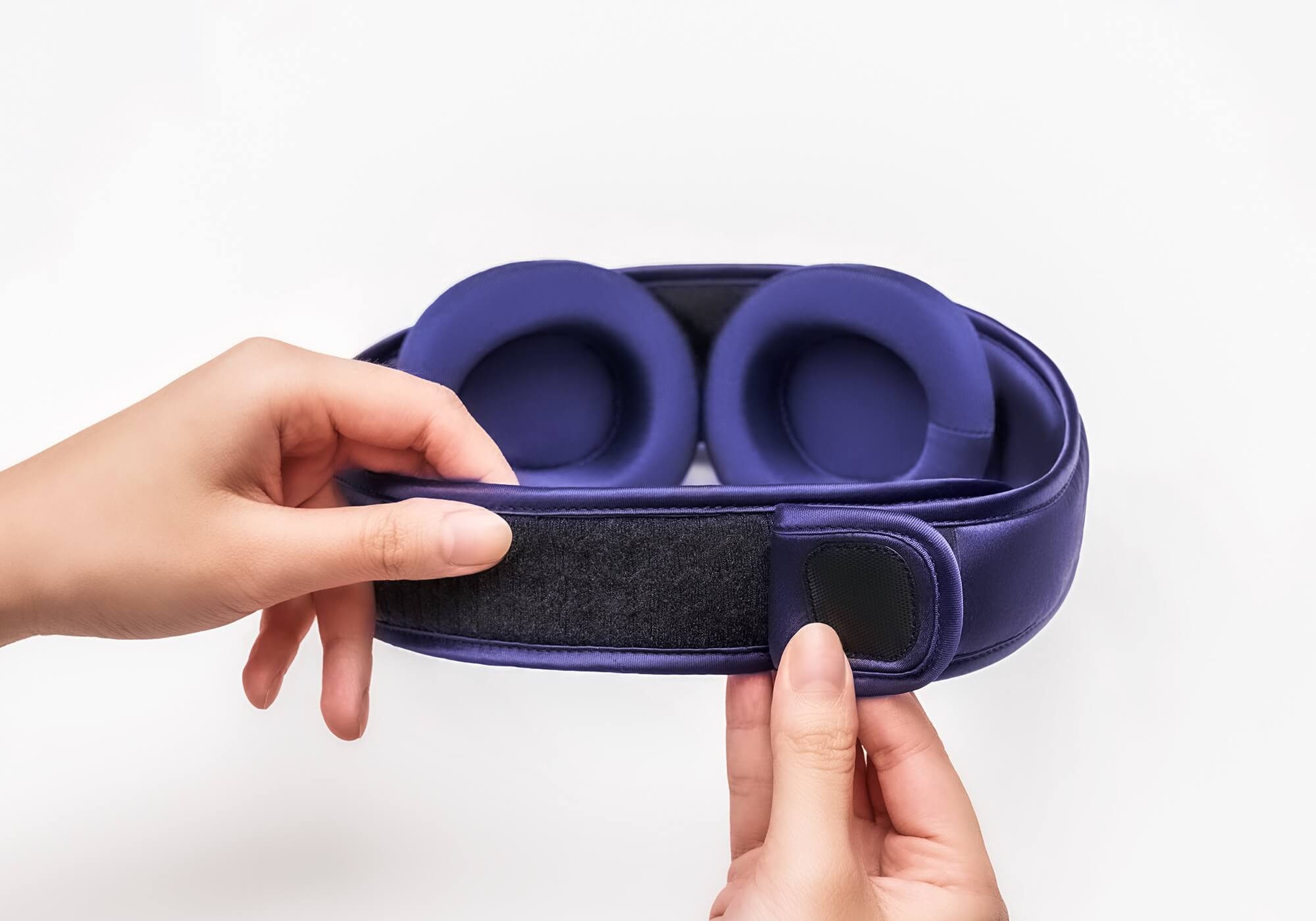 Two hands adjusting the hook-and-loop back closure of a purple silk sleep mask, revealing two adjustable, padded eye cups.