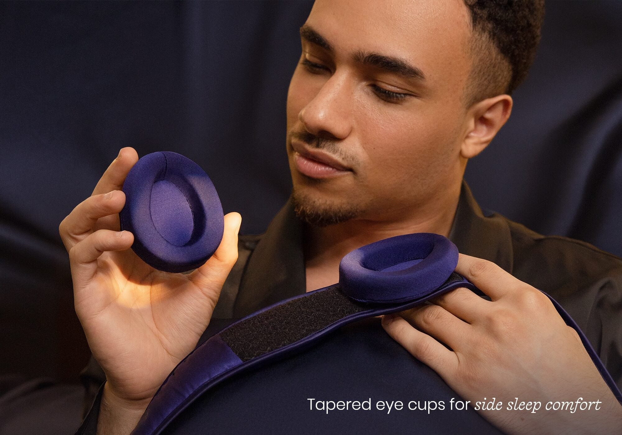 A man examines a detachable, purple silk eye cup from a sleep mask. Captioned "Tapered eye cups for side sleep comfort".