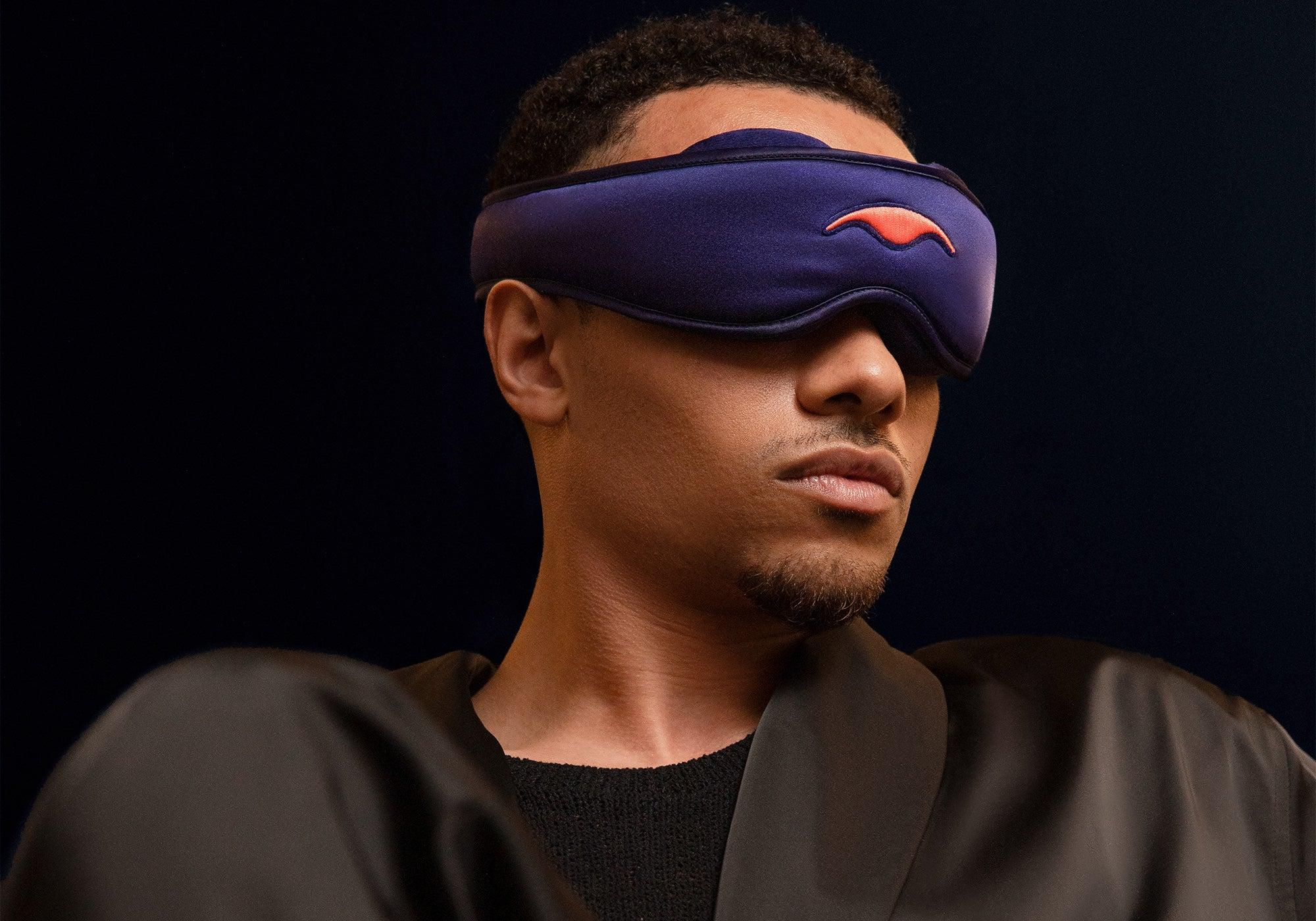 Low-angle studio profile shot of a man wearing a purple Manta Sleep Mask SILK over his eyes, paired with a black silk robe.