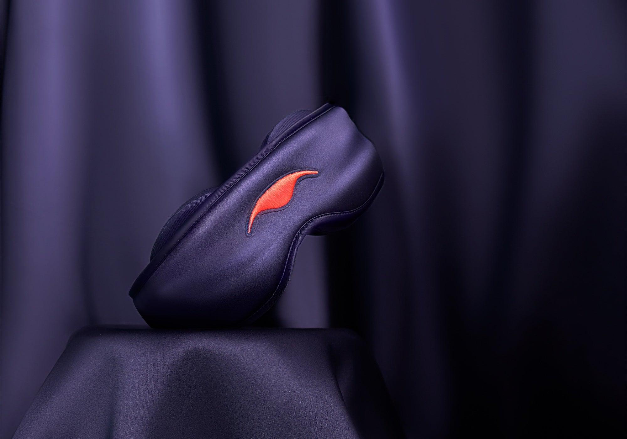 A purple silk sleep mask balancing elegantly on a pedestal draped in matching lustrous purple fabric.