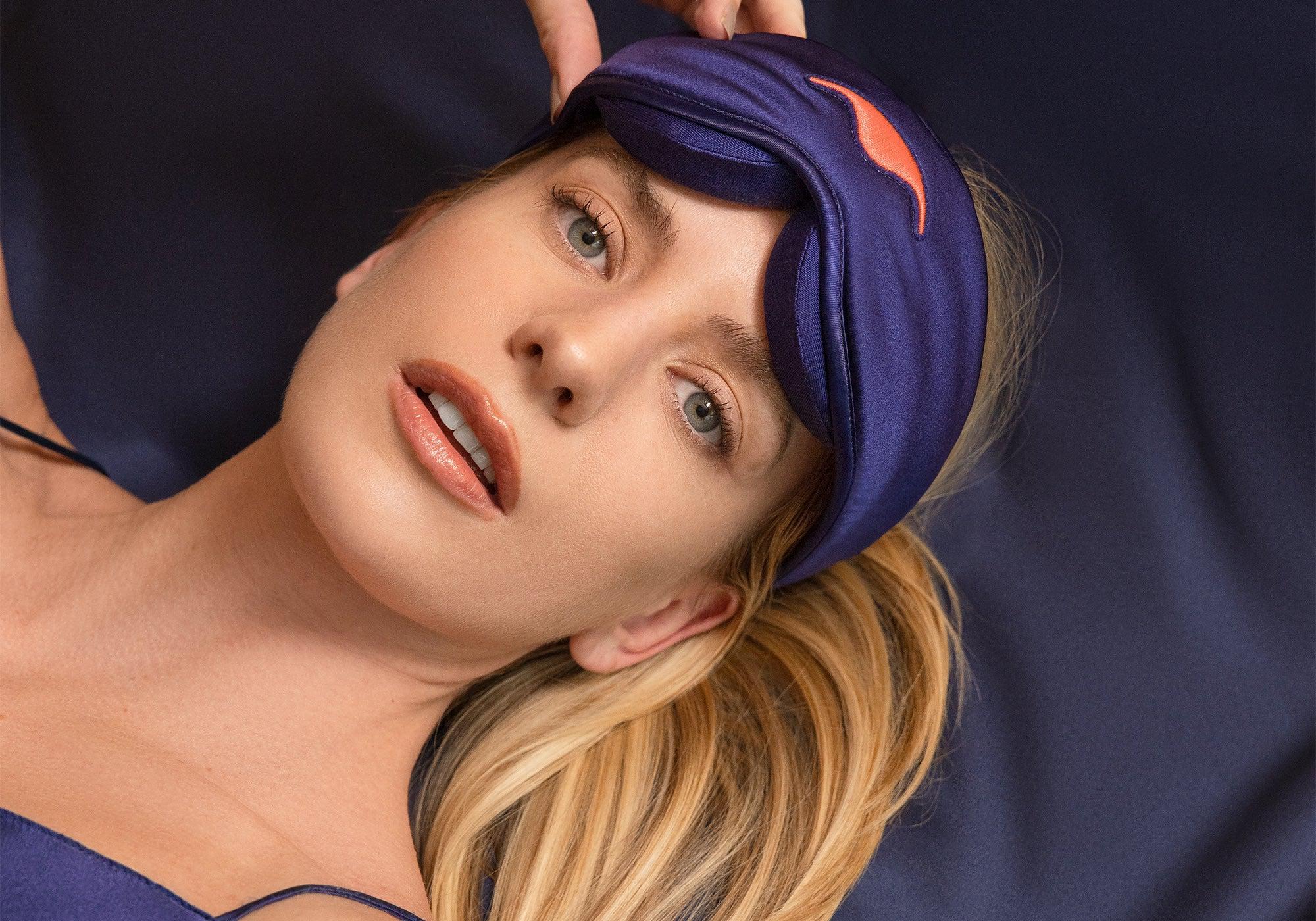 A woman with blonde hair lying down on a dark blue background, lifting a purple silk sleep mask up onto her forehead.