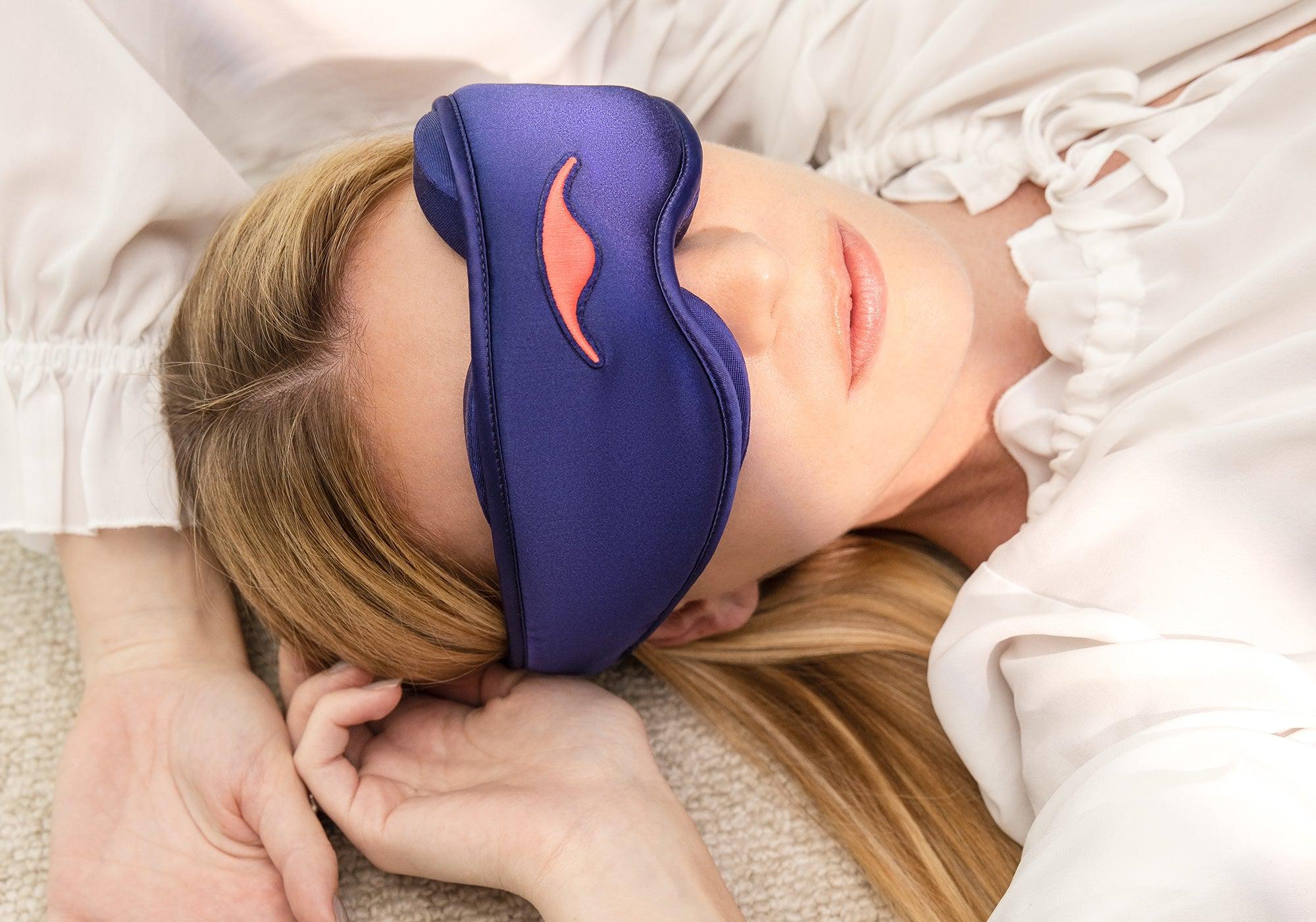 Close-up of a blonde woman lying down peacefully while wearing a purple silk sleep mask with an embroidered coral logo.