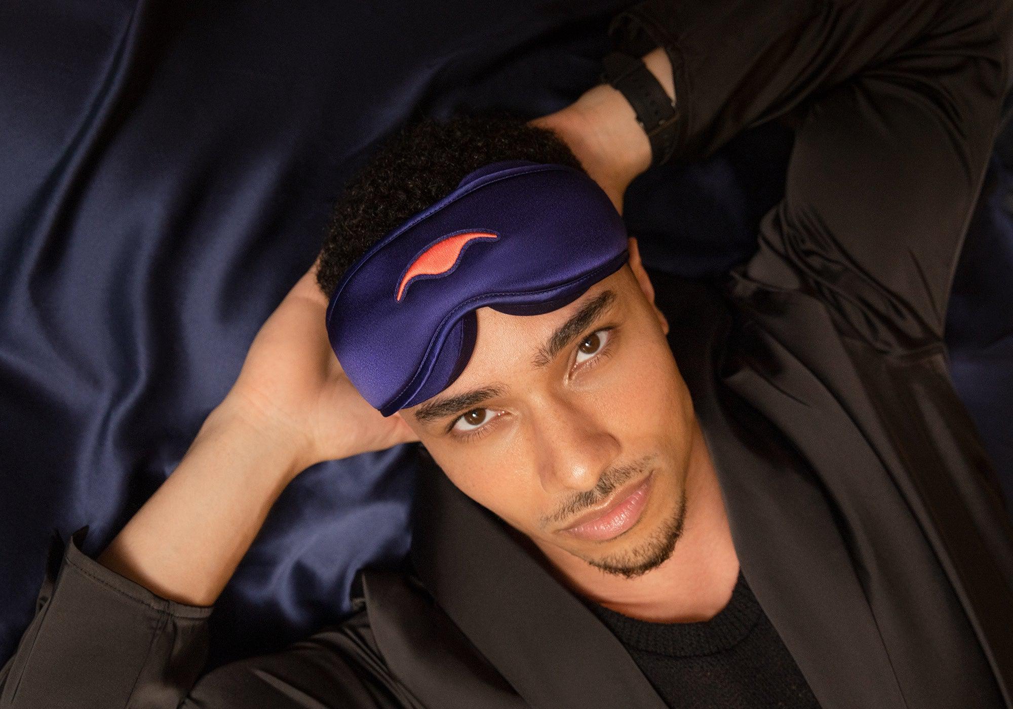 High-angle shot of a man lying down on dark silk sheets, wearing a purple Manta Sleep Mask SILK propped up on his forehead.