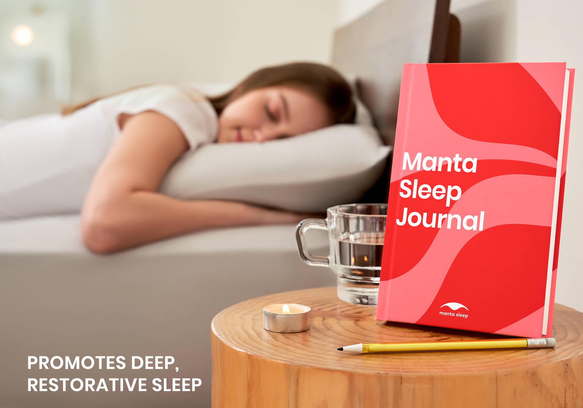 A red Manta Sleep Journal on a wooden table next to a candle and a glass of water, with a woman sleeping out of focus in the background.