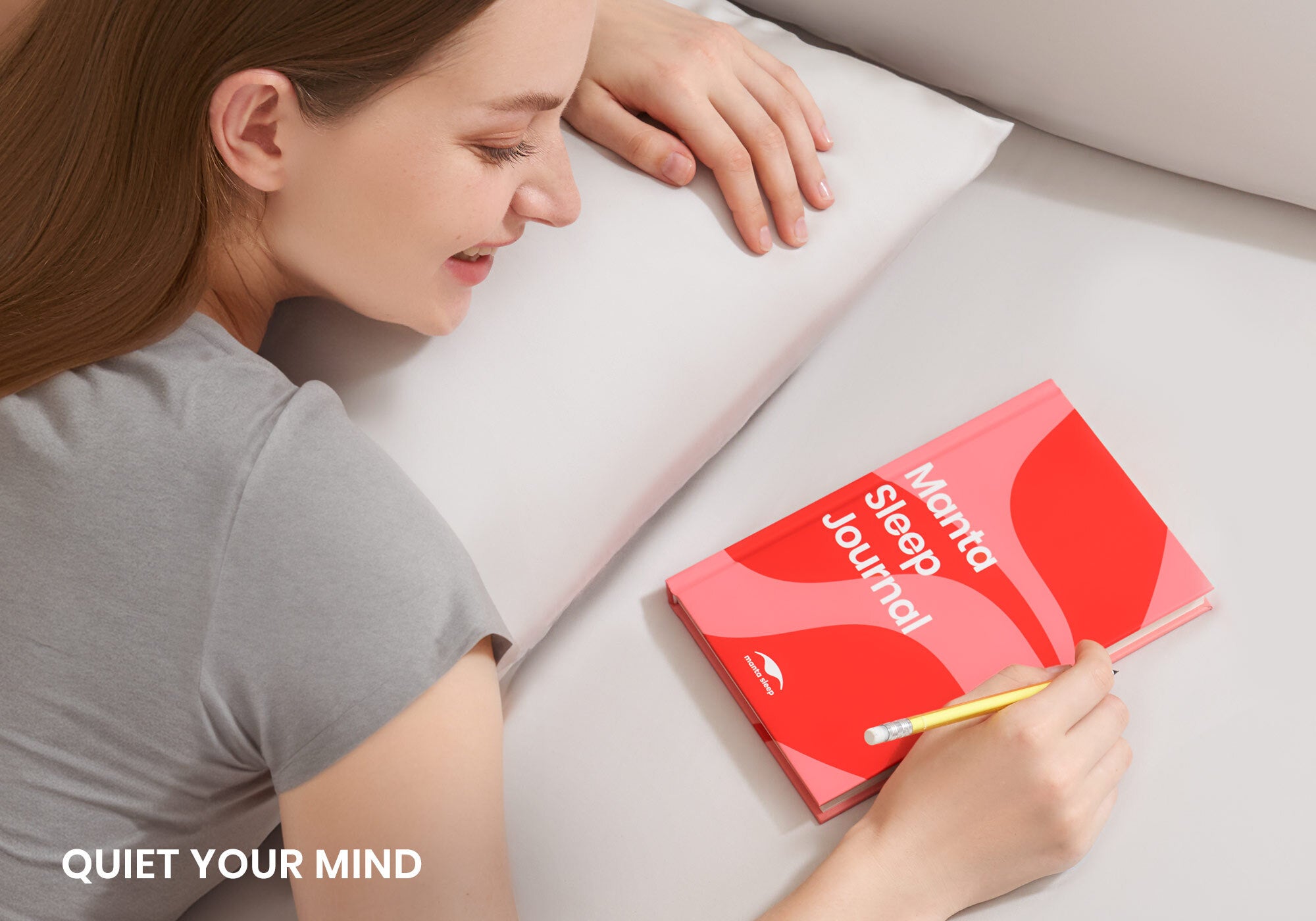 A smiling woman resting her head on a white pillow next to a red Manta Sleep Journal on the bed. Overlay text reads "Quiet your mind".