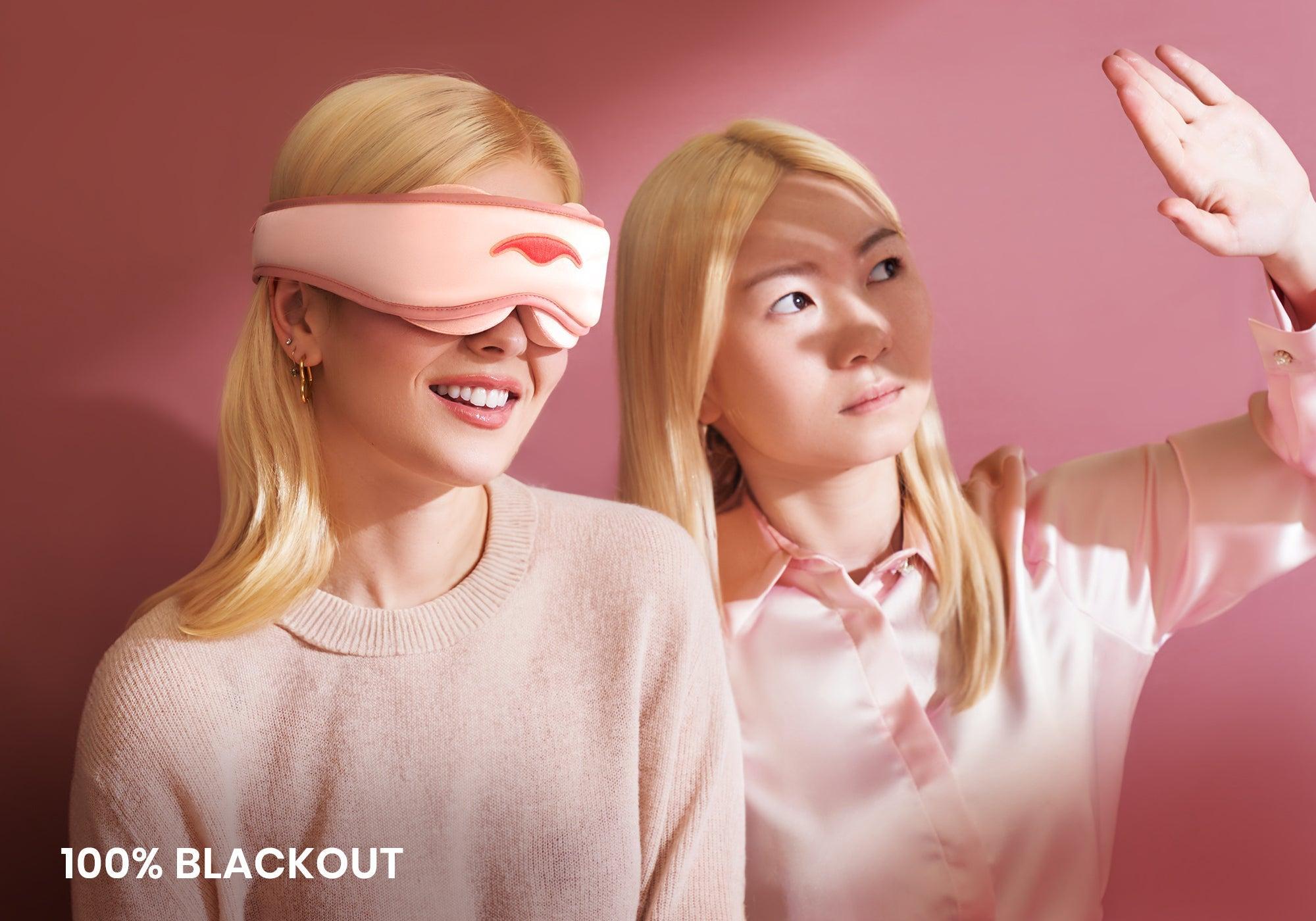 A woman shields her eyes from bright light while other woman rests beside her wearing a pink silk sleep mask, captioned "100% blackout".