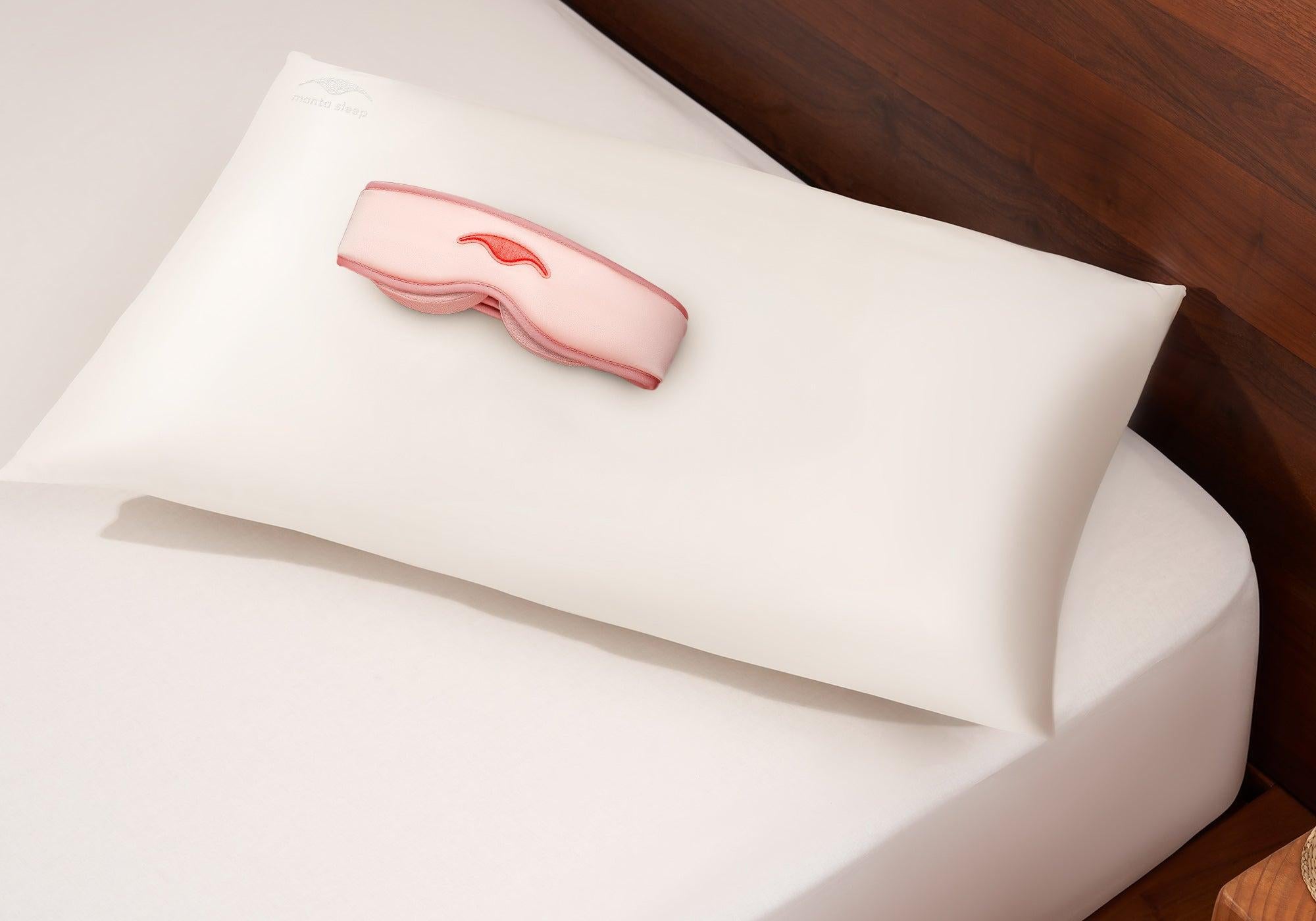 Sunset Pink Manta SILK Sleep Mask and pearl white Manta Silk Pillowcase stacked on a white bedsheet on a wooden bed.