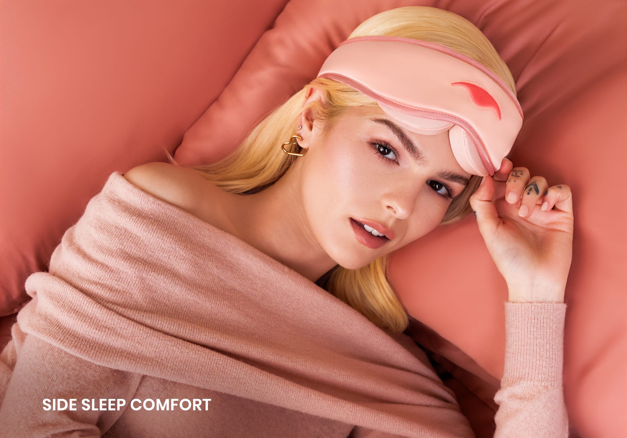 Top-down view of a blonde person lying on pink silk pillows, wearing a pink Manta SILK Sleep Mask pushed up on their forehead, with text overlay: "Side sleep comfort".