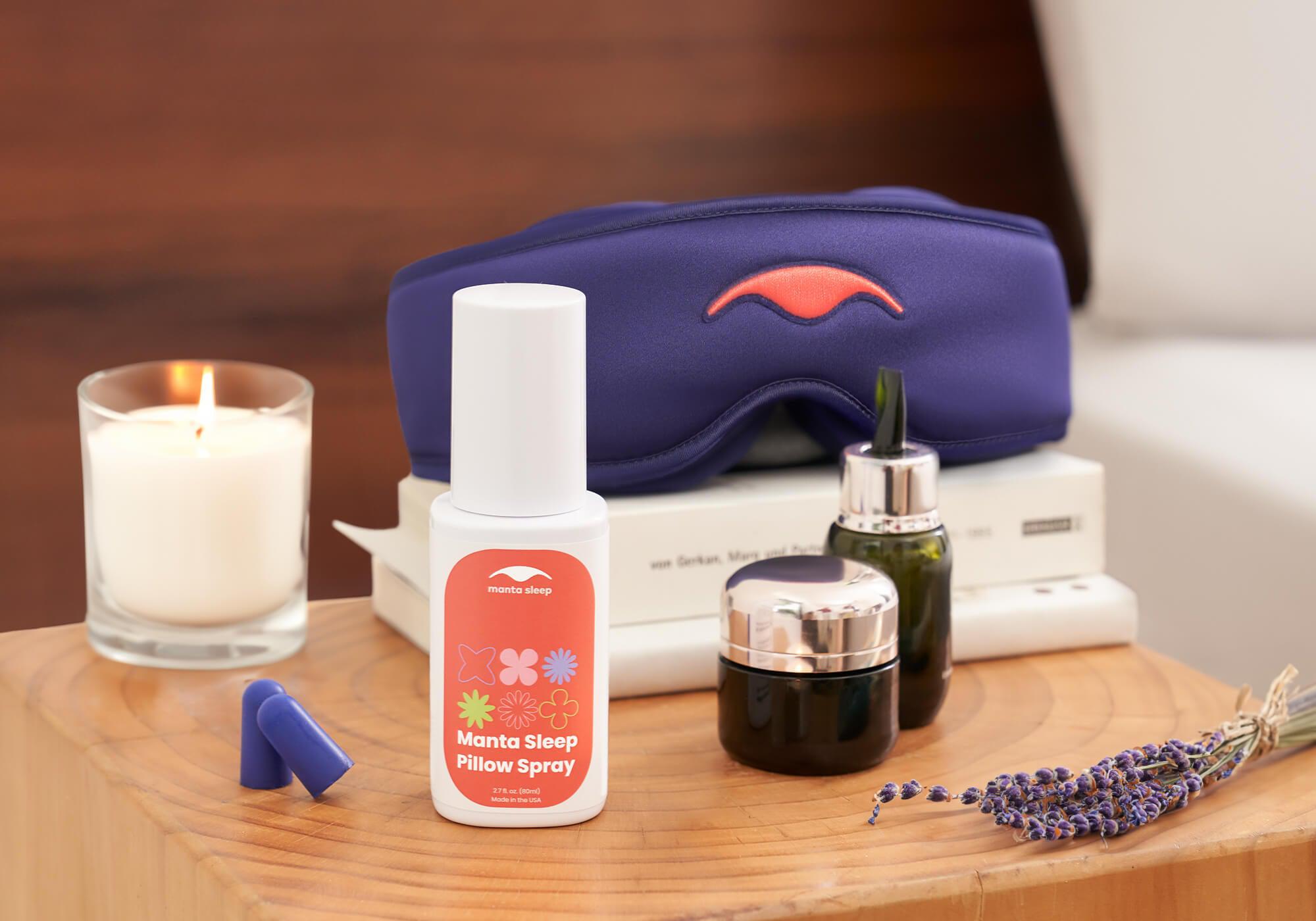 A nightstand with purple Manta SILK Sleep Mask on top of books, next to a burning candle, essential oils, and Manta Sleep Pillow Spray in the foreground.