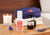 A nightstand with purple Manta SILK Sleep Mask on top of books, next to a burning candle, essential oils, and Manta Sleep Pillow Spray in the foreground.