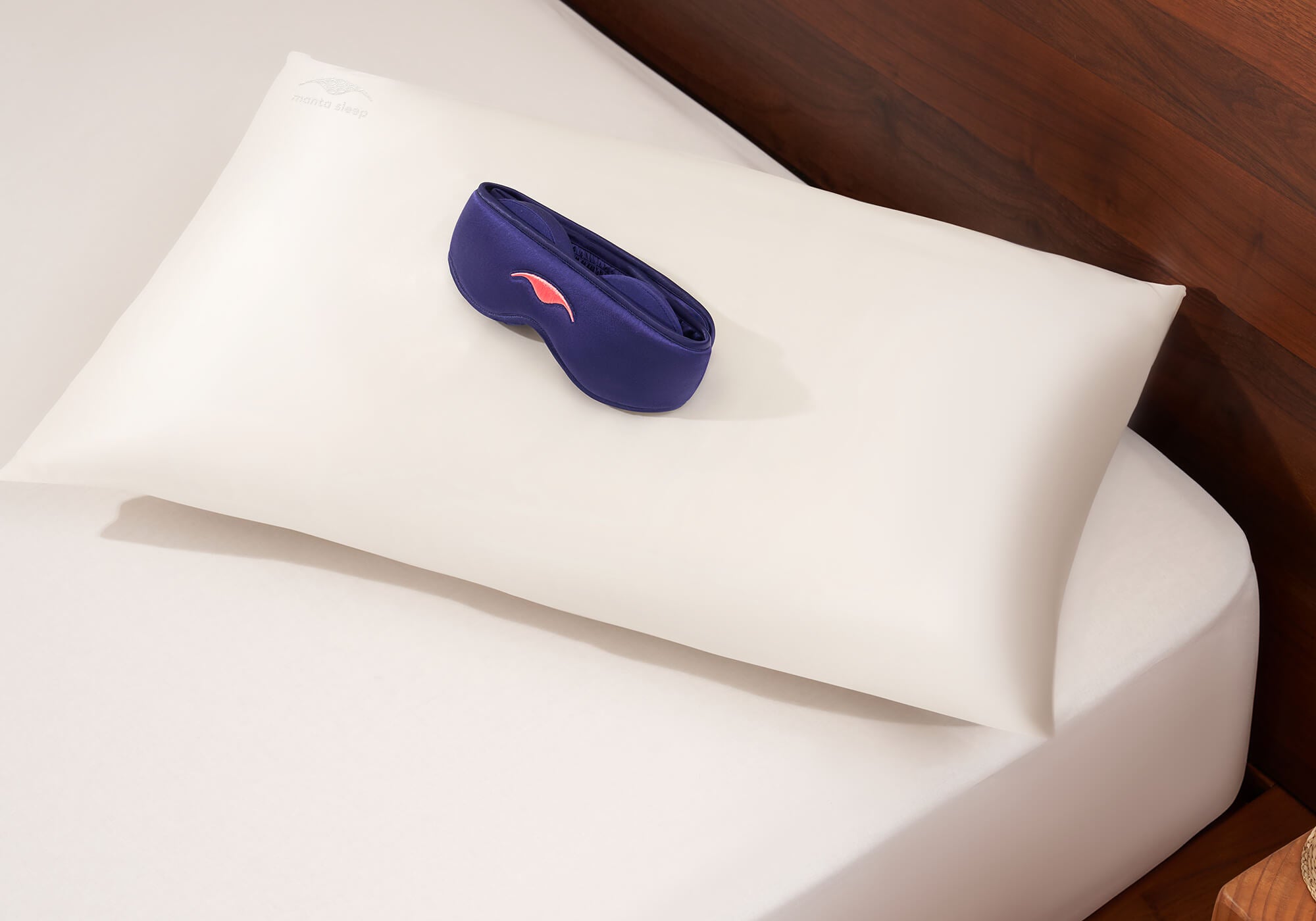 Purple Manta SILK Sleep Mask and pearl white Manta Silk Pillowcase stacked on a white bedsheet on a wooden bed.