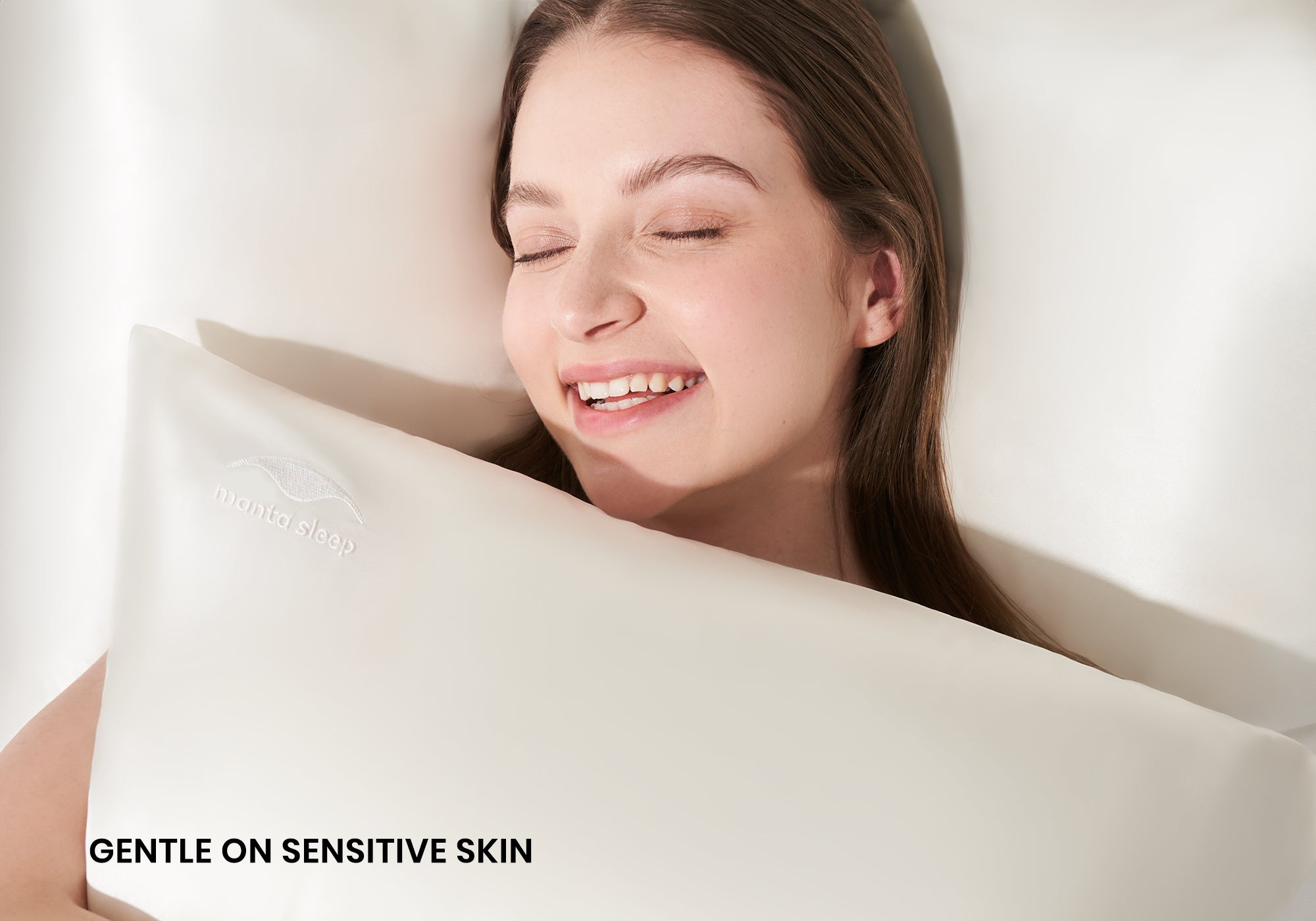 A smiling woman with closed eyes hugging a pearl white Manta Sleep silk pillowcase. Text overlay: "Gentle on sensitive skin".