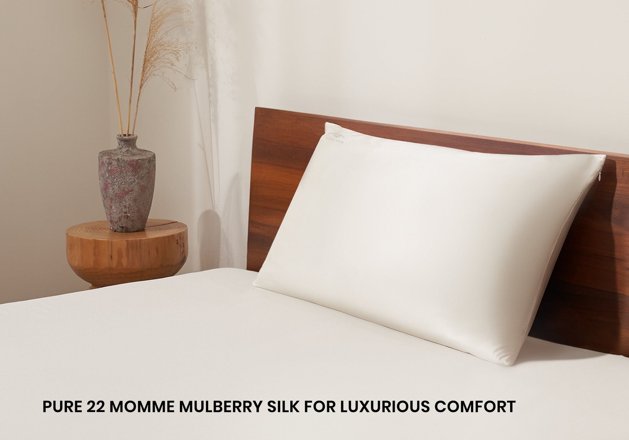 A pearl white Manta Sleep silk pillowcase on a bed next to a wooden side table with a vase. Text overlay: "Pure 22 momme mulberry silk for luxurious comfort".