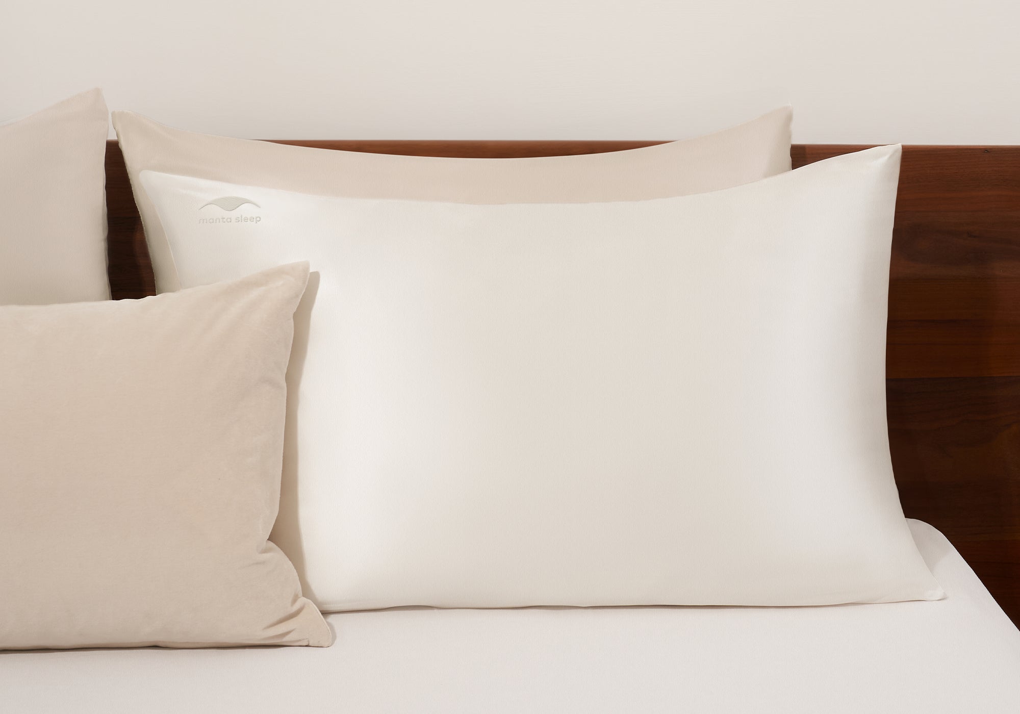 A pearl white Manta Sleep silk pillowcase arranged on a bed with matching neutral-toned accent pillows against a wooden headboard.