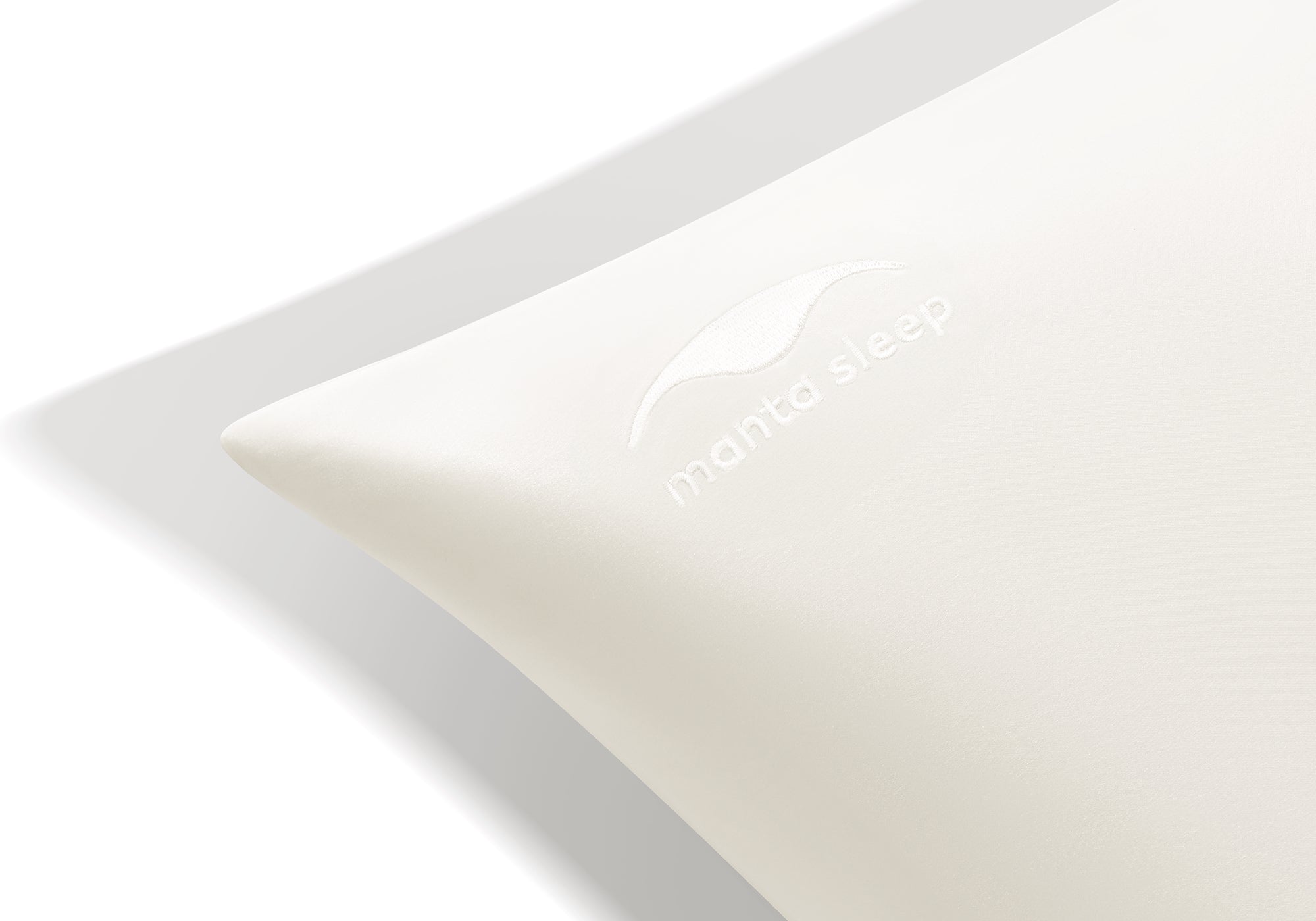 Extreme close-up of the tonal, embroidered Manta Sleep logo on the corner of a pearl white silk pillowcase.