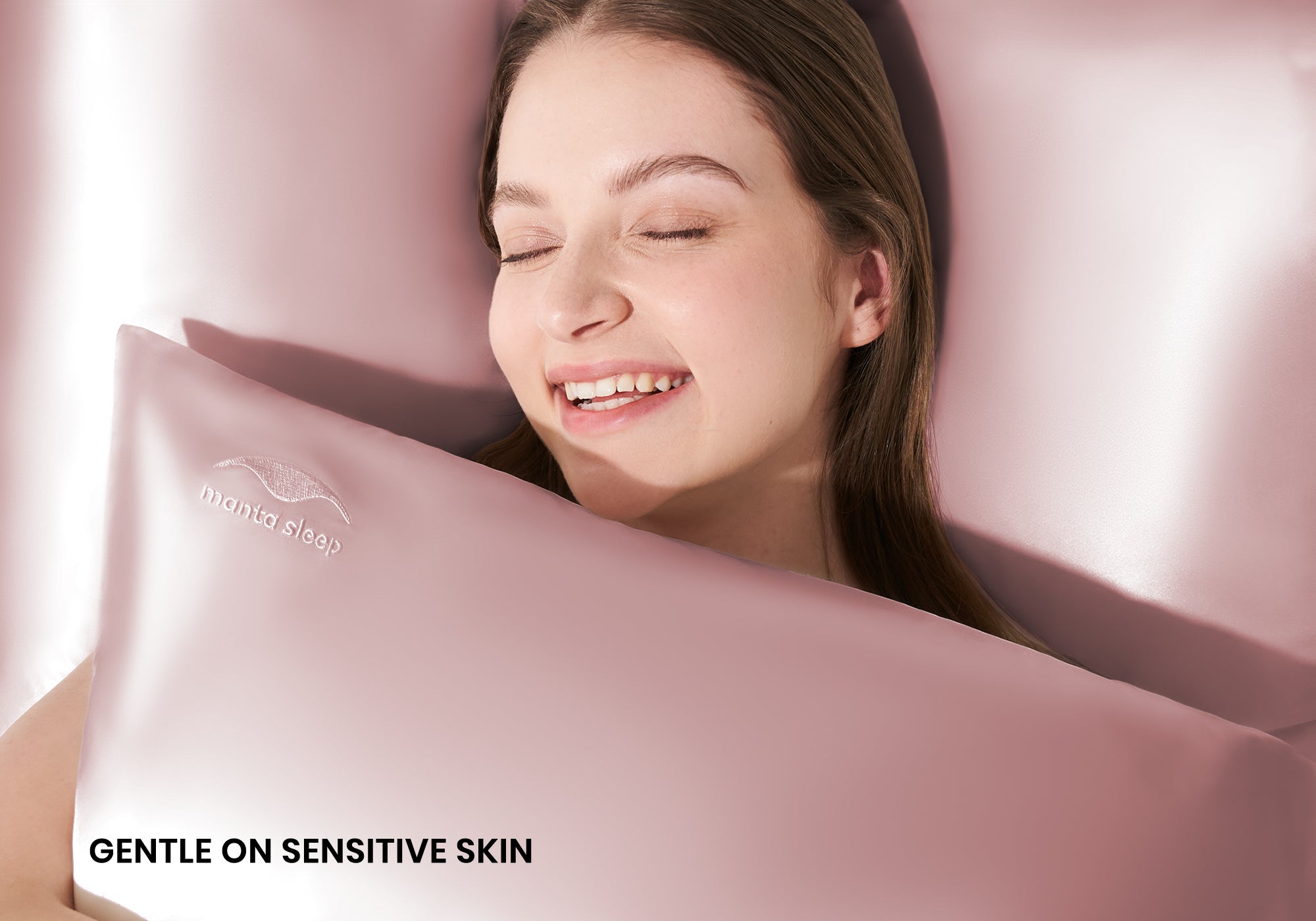 A smiling woman with closed eyes hugging a rose pink Manta Sleep silk pillowcase. Text overlay: "Gentle on sensitive skin".